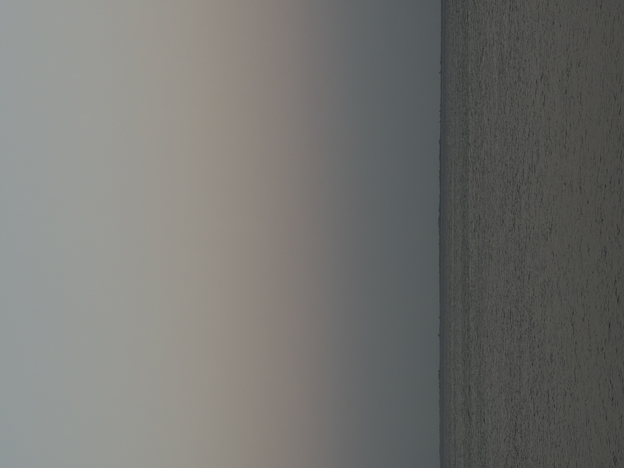 The image depicts a vast expanse of water under a pale, hazy sky. The horizon line is subtle, dividing the muted grey water from the gradient of pale blues and greys in the sky. There are no prominent objects or subjects visible in the foreground or midground. In the far distance, along the horizon, a faint, dark line suggests a distant landmass or a cluster of low-lying structures, possibly buildings or trees. The water appears calm with subtle ripples, reflecting the diffused light of the sky. The overall impression is one of quiet expanse and muted tones, characteristic of twilight or an overcast day. The location is the Volga River in Kamyshin, Russia.