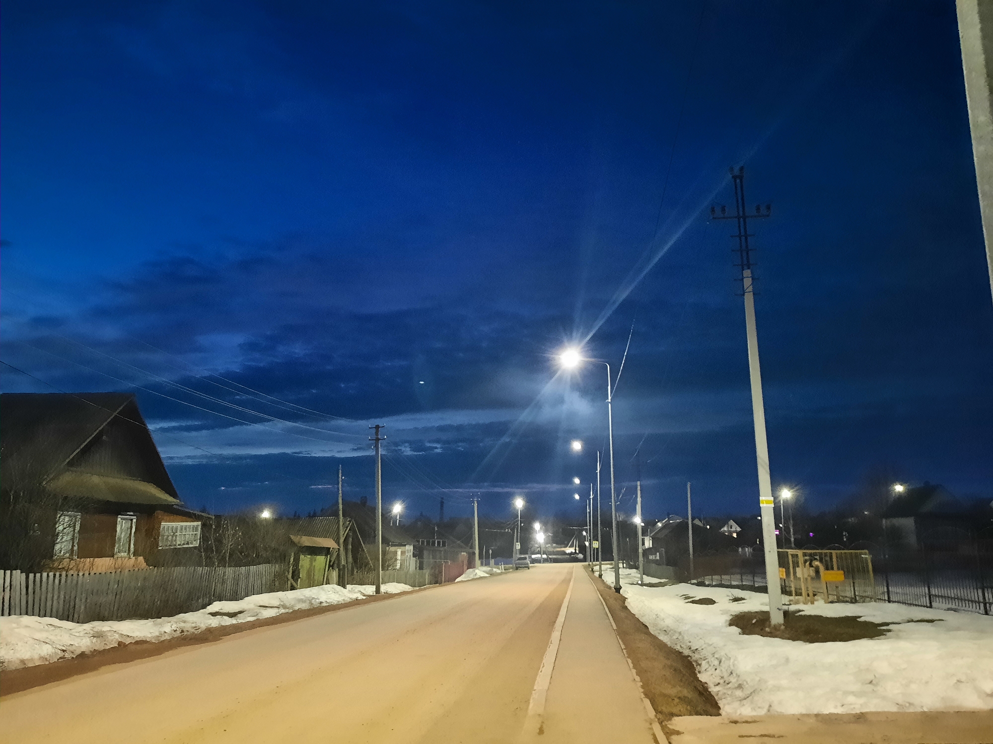 The image depicts a street in Krasnogorskoye, Russia, at dusk or night. The sky is a deep blue, with some cloud cover visible. Streetlights illuminate the road and surrounding areas. Snow is present along the sides of the road and in yards. The road itself appears to be paved and is free of traffic. Houses and fences line both sides of the street. Power lines are visible, supported by poles. A small playground is visible to the right side.