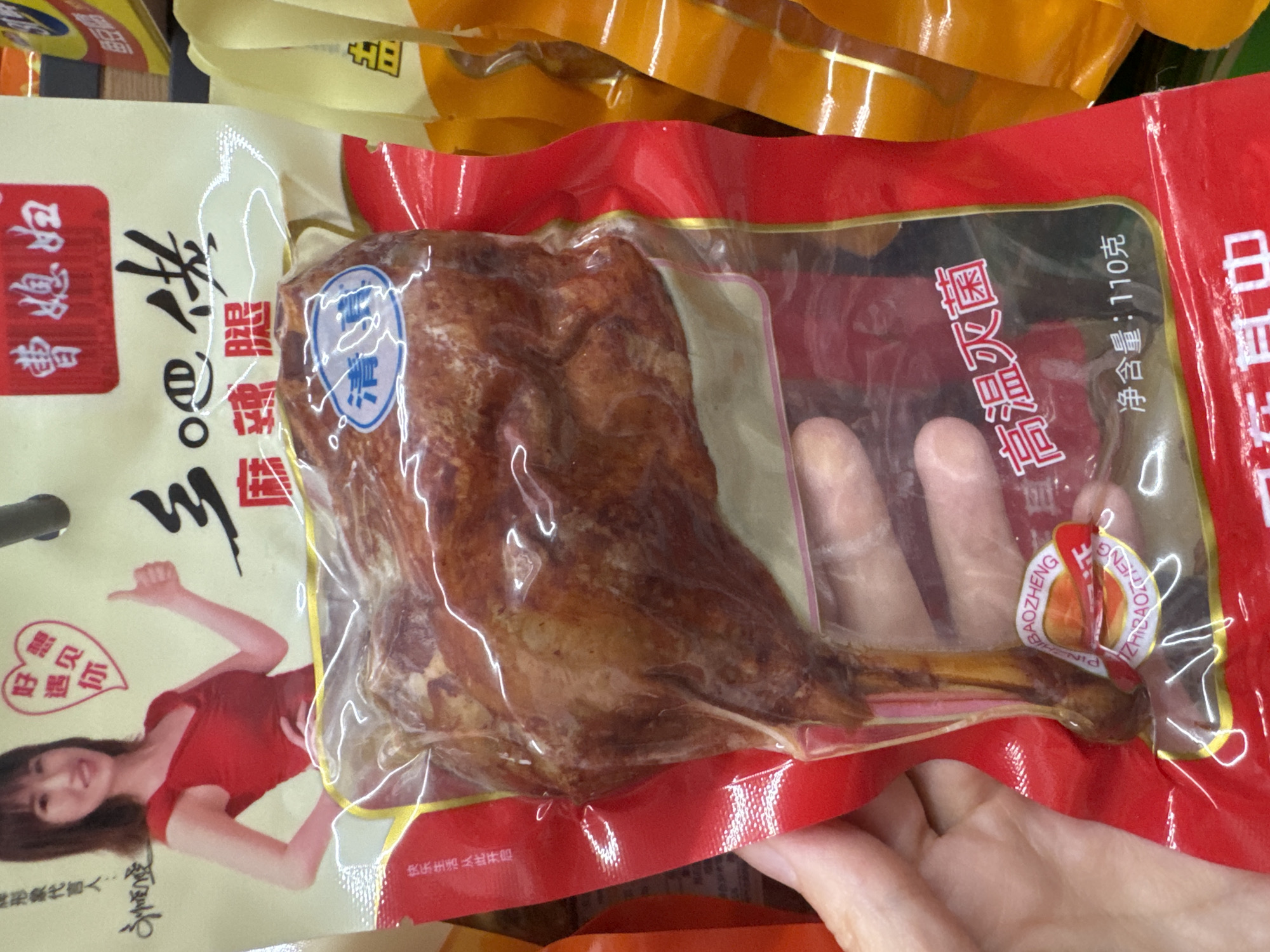 This image, observed in Wulingyuan, China, depicts a light-skinned hand holding a vacuum-sealed package of a processed poultry leg. The product itself is a reddish-brown, cooked poultry leg (likely chicken or duck), visible through clear plastic. A small blue circular label with the white Chinese characters "清真" (Halal) is affixed to the clear vacuum seal.
The main product packaging is predominantly red and white. The red section, visible on the left and bottom, displays white Chinese characters, including "高温灭菌" (High-temperature sterilization) and "净含量: 110克" (Net weight: 110 grams). The white section, at the top and upper right, features a graphic of a smiling woman with dark hair, wearing a red top, giving a thumbs-up gesture. Above her, a red heart graphic contains the text "好想遇见你" (So eager to meet you). Other Chinese characters on the white background include "曹媳妇" (Cao Xifu / Wife Cao) and "麻辣腿" (Spicy Leg).
The background shows out-of-focus yellow and orange packaging from other products, consistent with a retail store environment. No direct interactions beyond the hand holding the product are visible.