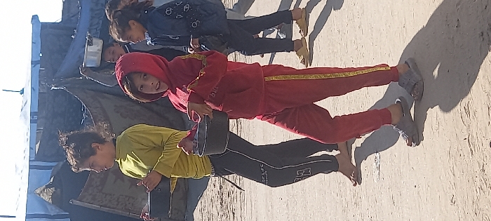 The image captures a group of children outdoors in Khan Yunis, Palestinian Territory, under bright sunlight, with makeshift shelters visible in the background. In the immediate foreground, a boy with dark, disheveled hair, wearing a lime green long-sleeved shirt and dark track pants with white lettering, is barefoot. He holds a dark-colored pot in both hands and looks towards the left. To his right, a younger boy, wearing a red hooded tracksuit with yellow vertical stripes along the arms and legs, and grey slip-on sandals, also holds a dark-colored pot in both hands. This boy is looking directly at the viewer with a slight smile. In the background, partially obscured, are several other children. One child has a light-colored metallic pot balanced on their head. Another child is visible wearing a dark blue jacket with white patterns and light-colored slip-on shoes. The ground is unpaved earth, casting distinct shadows from the figures. The background features structures constructed from blue and dark-colored tarpaulins and fabrics, indicative of temporary dwellings.