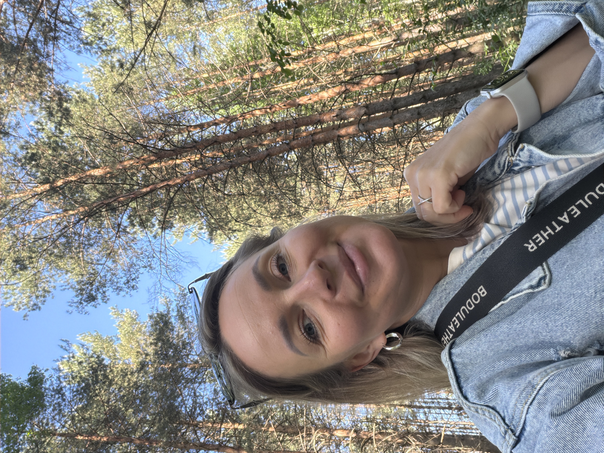 A selfie image shows a woman with light skin, blonde hair, and blue eyes, wearing a denim jacket, a striped shirt, and a silver ring. The background features a dense forest canopy with visible blue sky peeking through. The woman is outdoors, possibly in a forest or park-like setting near Chaikovsky, Russia.