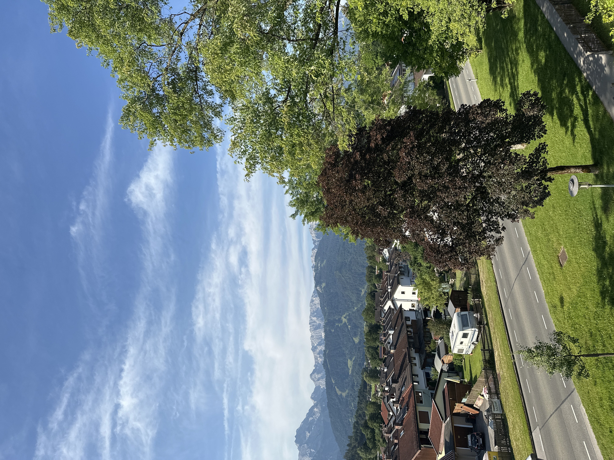 The image shows a view of Garmisch-Partenkirchen, Germany, on a sunny day. In the foreground, a paved road runs horizontally across the frame, bordered by a grassy lawn on the right and a fence and more grass on the left. Several trees are present, including a large dark-leafed tree in the middle ground and a lush green tree on the right. Beyond the road and lawn, there is a residential area with houses featuring terracotta roofs. Further in the background, a vast mountain range dominates the skyline, with its peaks partially obscured by clouds. The sky is blue with scattered white clouds. There are no people visible in the image.