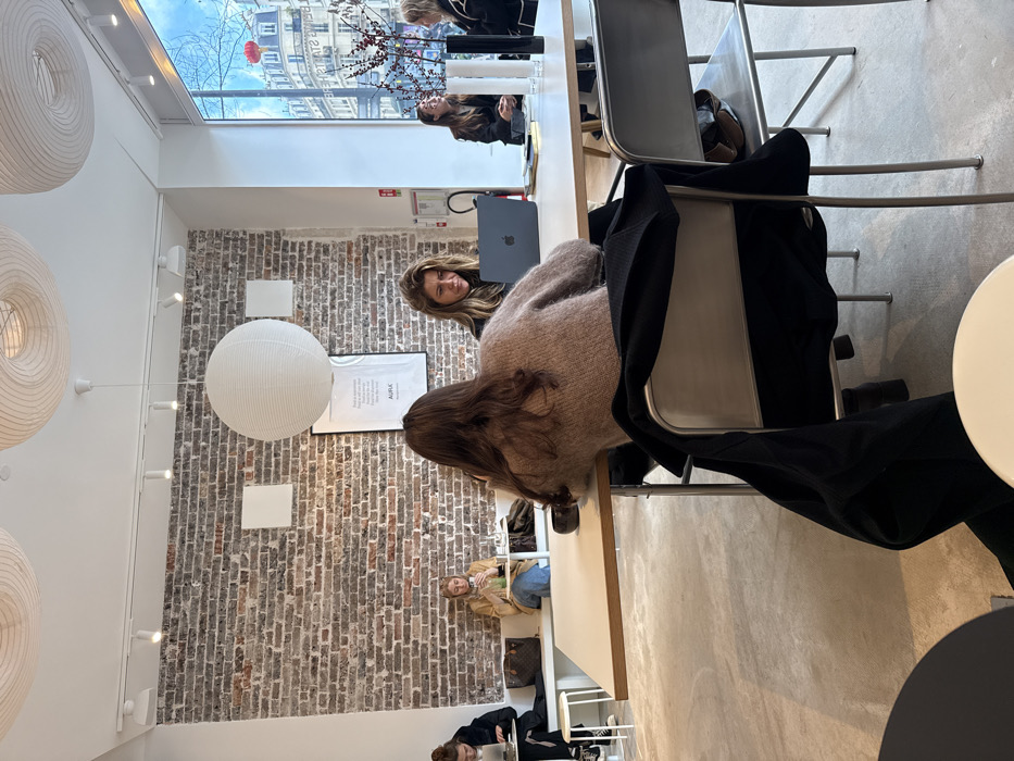 The image depicts an interior view of a modern cafe or co-working space located on Rue Delavigne in Paris, France, identifiable by visible street signage and typical Parisian architecture seen through a large window. The space features an exposed brick wall along the left and rear, juxtaposed with white painted walls and a light grey, possibly concrete, floor. The ceiling is white, fitted with track lighting and three large, spherical white paper lanterns.
In the foreground and mid-ground, a person with long, wavy brown hair, wearing a light brown textured sweater, is seated with their back to the camera at a rectangular light-colored table. A black jacket is draped over the back of their silver-framed chair. Directly opposite, a woman with wavy brown hair and a dark top is focused on a silver laptop.
To the left, against the brick wall, a long white bench serves as seating. Two individuals occupy this bench at separate small white tables. The furthest individual, wearing dark clothing, uses a laptop. Closer, a blonde woman in a light brown jacket and blue jeans holds a green beverage in a clear glass; a brown monogrammed bag rests beside her on the bench.
In the background on the right, two women are seated near the expansive window. One woman with dark hair is engaged in conversation with the other, who is partially obscured. Their table holds a dark vase with red-berried branches and several white cylindrical decorative items.
Through the window, classic Parisian Haussmannian buildings with light stone facades, ornamental balconies, and grey mansard roofs are visible under a blue sky. A red spherical lantern hangs outside. A black-framed poster, partially legible with the word "AURIC," is mounted on the brick wall.
