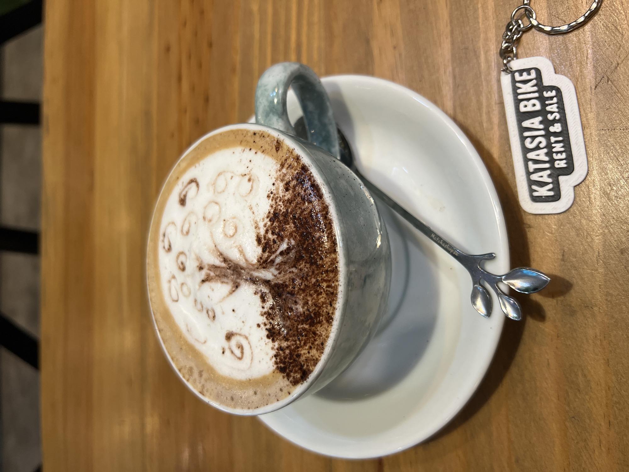 A bird's-eye-view photograph reveals a coffee cup with latte art, placed on a saucer with a small spoon, beside a keychain that reads "KATASIA BIKE RENT & SALE." The location, inferred from the keychain, is Nha Trang Ward, Vietnam. The coffee cup is approximately centered in the frame, filled with a latte exhibiting a textured design made from coffee granules or powder. The spoon, with a decorative leaf design, rests on the white saucer. The keychain is positioned to the right of the saucer on a wooden table. The image suggests a café or coffee shop setting. There are no people visible. The lighting appears to be natural or indoor lighting with diffused brightness. The overall composition suggests a promotional or product shot.
