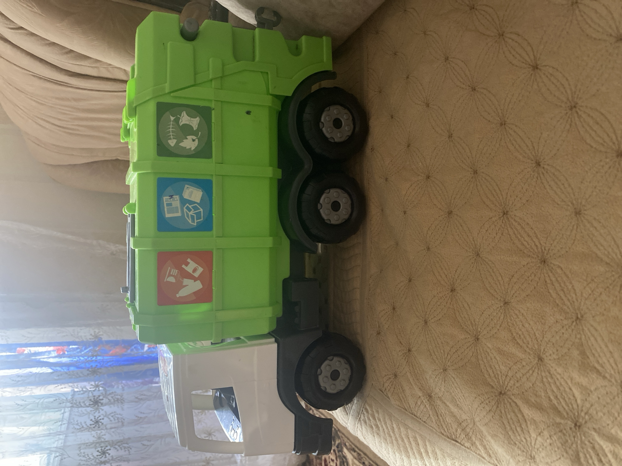 A toy garbage truck is positioned on a beige quilted surface. The truck is primarily green and white, with three colored recycling symbols on its side: one red with a bottle and plastic bag, one blue with a box and paper, and one green with a fish skeleton, leaf, and apple core. The truck has black wheels. In the background, sheer curtains with a floral pattern are visible, allowing light to filter through. The overall setting appears to be indoors, possibly a home environment. There is no indication of the city of Kizilyurt, Russia in the image.