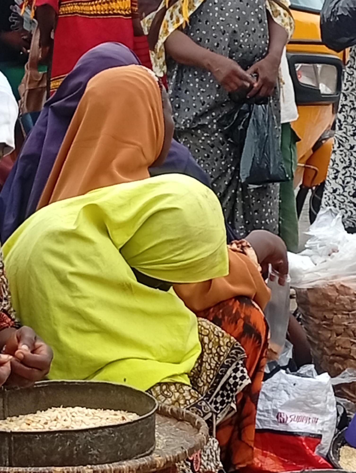 In Maiburiji, Nigeria, a group of individuals is gathered outdoors. At the forefront, a woman wearing a green head covering is seated, occupied with sorting through what appears to be grain from a metal container. Behind her, another woman is seen wearing an orange head covering over a purple garment. Further back, a woman dressed in a grey and white patterned dress holds a black plastic bag. A yellow vehicle is partially visible in the background. The scene is cluttered with various bags and materials.