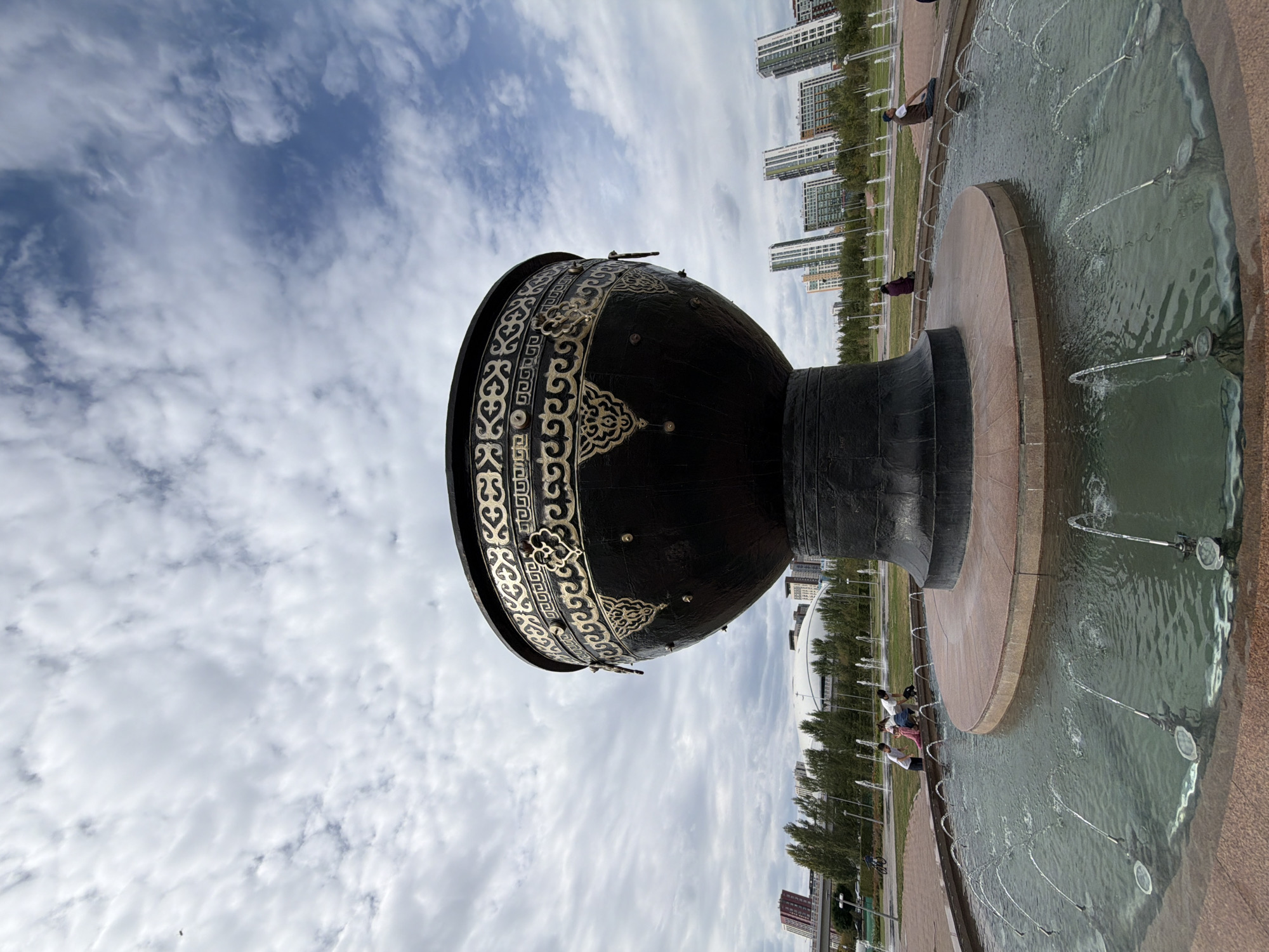 A low-angle, wide shot captures a large, black, ornate cauldron monument with gold detailing situated in a fountain in Astana, Kazakhstan. The monument, which appears to be made of metal, is supported by a dark, textured base and a reddish-brown circular platform. The fountain itself has several jets of water spraying upwards from within a wide, shallow basin. In the background, modern high-rise buildings with distinctive architecture are visible, along with trees and a white-domed structure that may be a stadium or exhibition center. The sky is cloudy, with patches of blue peeking through. In the mid-ground, a few people are scattered around the fountain, some sitting and observing, while a family with children and a dog are gathered near the edge of the fountain. Another individual is seen sitting on a bench to the right, and a person on a bicycle is in the far left background. The scene suggests a public park or plaza in a developed urban area.