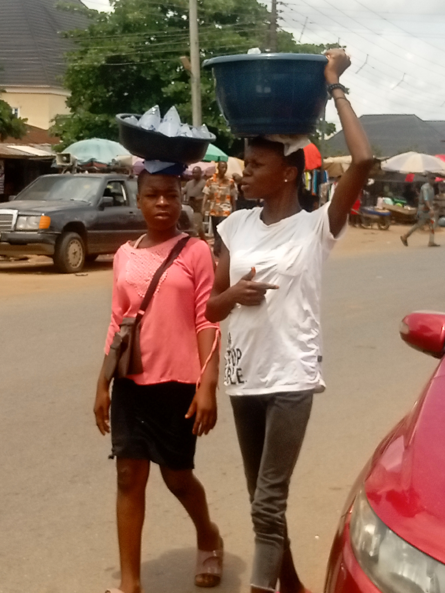 Two young women are walking on a paved road in Ogidi, Nigeria. The woman on the left wears a coral long-sleeved shirt, black skirt, and tan sandals, and carries a brown purse. She has a bowl of clear, triangular-shaped items balanced on her head. The woman on the right wears a white t-shirt, dark jeans, and no visible shoes. She has a dark blue bowl balanced on her head and is gesturing with her right hand. In the background, there is a gray SUV, various market stalls with umbrellas, and several people. The sky is overcast. A red car is partially visible on the right side of the image.