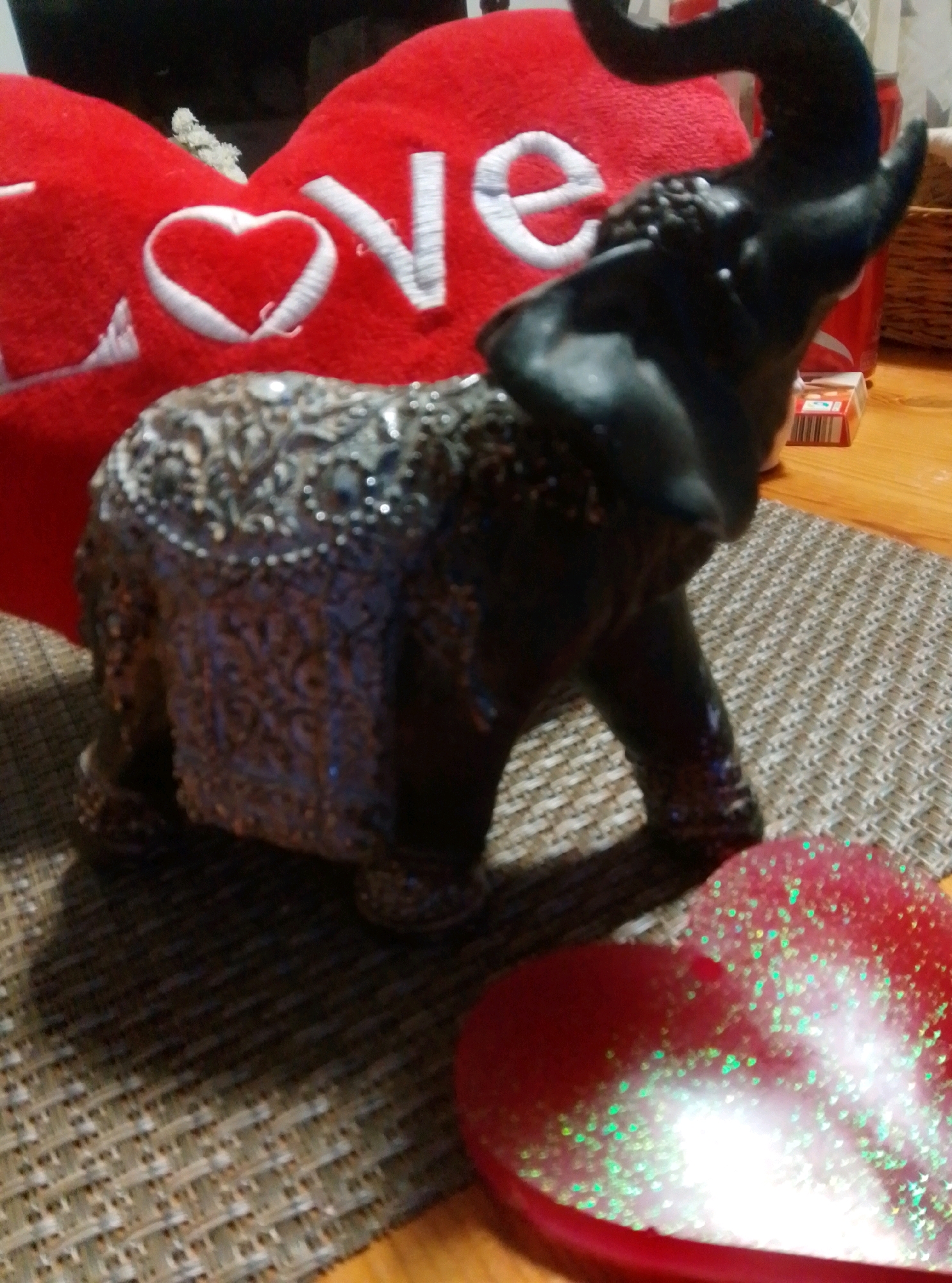 The image shows a close-up of a decorative elephant figurine placed on a woven placemat. The elephant is dark in color and intricately detailed with what appear to be raised patterns or embellishments. Behind the elephant, a red heart-shaped pillow with "LOVE" written in white lettering is visible. To the right of the elephant, a red and white box, possibly containing candy or a similar item, and a portion of a red can are also present. In the foreground, a red heart-shaped object with green glitter is partially visible. The overall setting appears to be a tabletop indoors. There is no indication of the city of Wedel, Germany in the image.