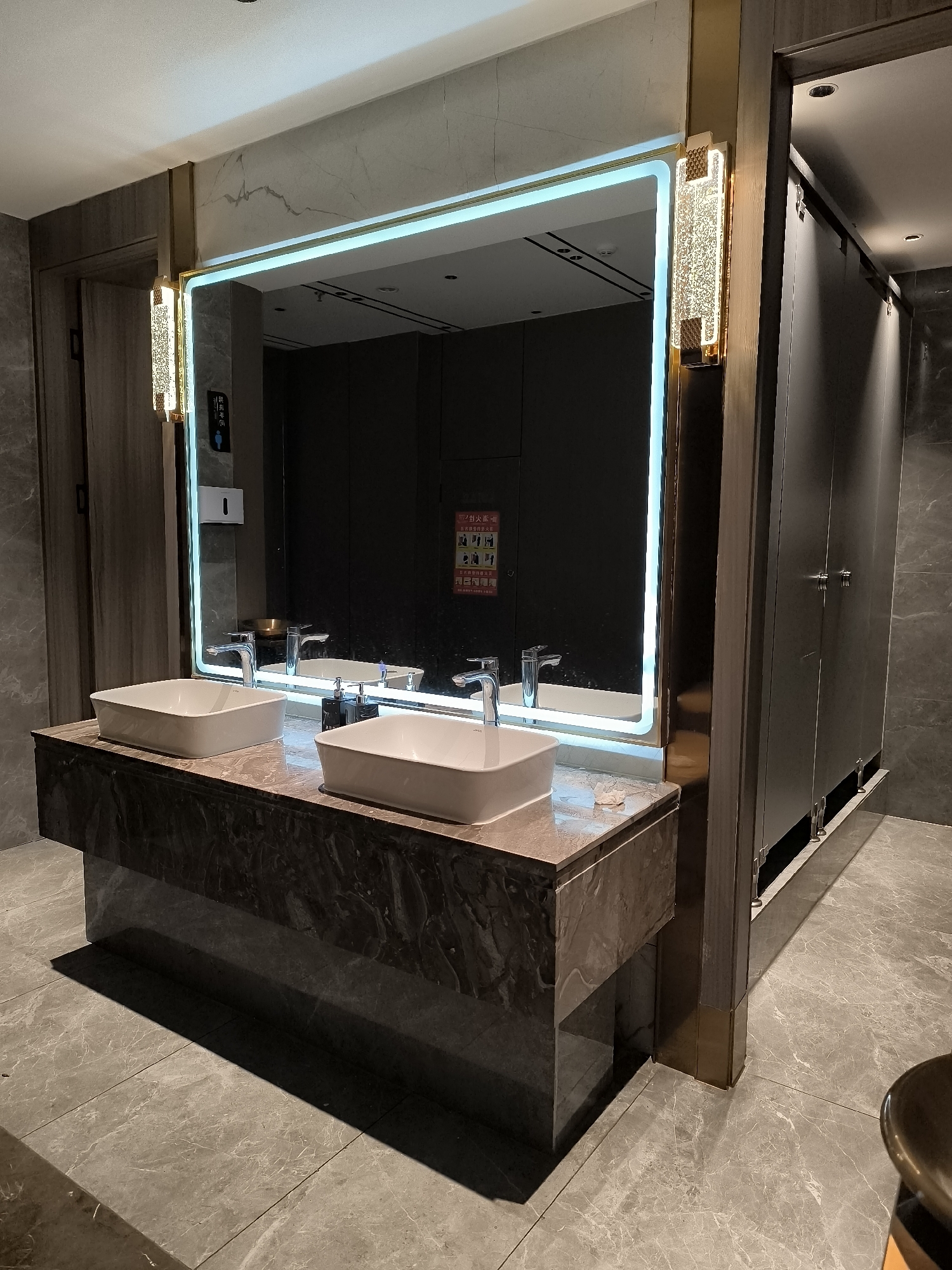 The image shows a modern public restroom, likely in the city of Ürümqi, China. The restroom features a large, well-lit mirror with LED lighting, reflecting two white ceramic sinks and chrome faucets. The vanity is made of marble with a dark, veined pattern. To the right of the vanity are several dark-colored stall doors, suggesting a multi-stall restroom. The flooring is tiled with a marble-like pattern in shades of grey. Decorative wall sconces with a textured glass design flank the mirror. A white soap dispenser is mounted on the wall to the left of the mirror, and a fire safety poster with pictograms is visible on the mirror itself. The overall impression is one of cleanliness and contemporary design.