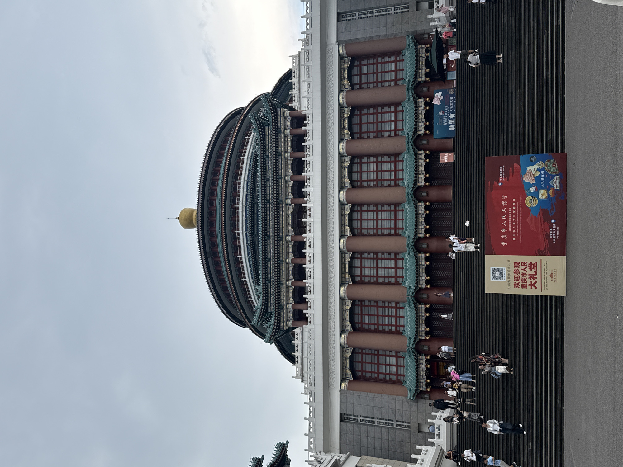 The image shows the Chongqing Great Hall of the People, a landmark building located in Yuzhong District, China. The grand edifice features a distinctive circular structure with multiple tiered roofs, adorned with traditional Chinese architectural elements such as upturned eaves and decorative brackets. A golden dome crowns the uppermost level. The building is fronted by a wide, imposing staircase, on which a diverse group of people are ascending and descending. Several individuals are captured in motion, walking, talking, or posing for photos. Some are dressed casually, while others appear to be tourists. Large banners displaying Chinese characters and illustrations are prominently placed on the steps, indicating an event or exhibition. The sky above is overcast, casting a soft light on the scene. The overall impression is one of a bustling public space centered around a significant cultural and architectural monument.