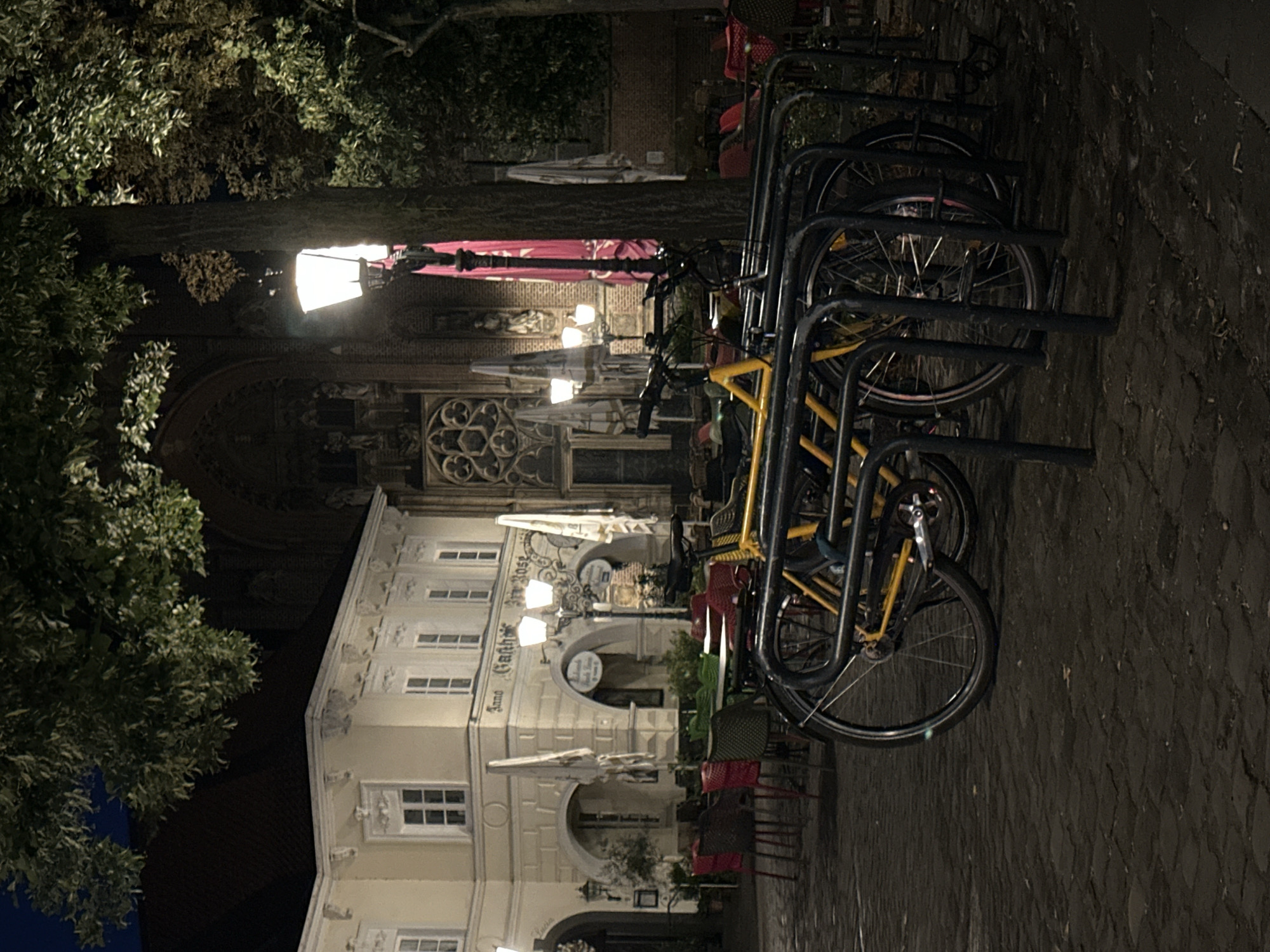 At night on a cobblestone plaza in Krefeld, Germany, several bicycles are parked in a rack in the foreground. The bicycles are dark-colored with some yellow accents. Behind the bicycles, an outdoor seating area for a restaurant is visible with tables and chairs. One building, possibly a historic gasthof, has architectural details such as arched doorways and windows. A lamppost illuminates the scene. In the background, a large, Gothic-style building with intricate stonework and a prominent rose window can be seen. Trees with foliage overhang the plaza.
