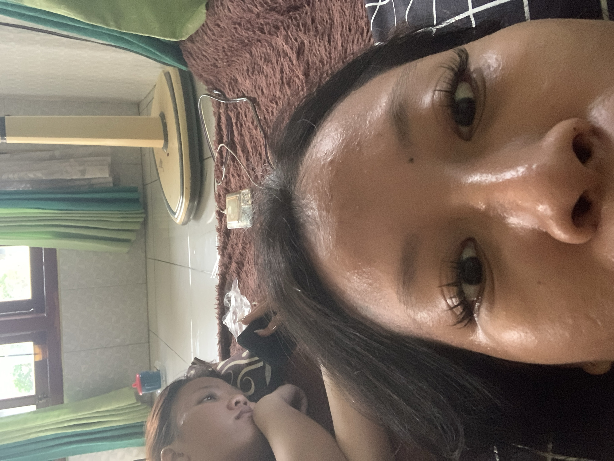 Two people are lying on a brown furry rug in what appears to be a residential interior. The person in the foreground is facing the camera, with their face taking up most of the frame. Their eyes are wide open, and they have long, dark eyelashes. The person in the background is lying on their side, with their head resting on a pillow. They are looking away from the camera. A floor fan is visible in the background, along with green curtains and a window. There is also a small blue cup with a red lid on the floor. The scene suggests a casual, indoor setting. It's not possible to determine the exact location in Jajag, Indonesia based solely on the visual information provided.