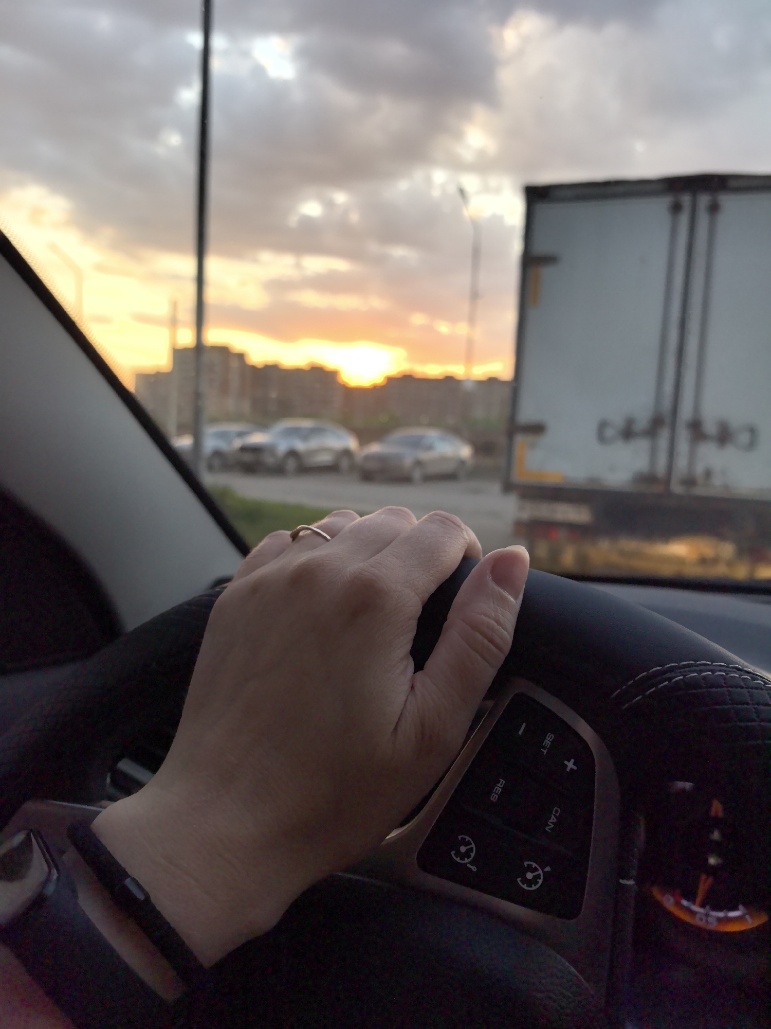 A photograph taken from inside a car depicts a person's hand on the steering wheel. The hand has a ring on the ring finger. The dashboard is visible, featuring buttons and a speedometer. Outside the car, the view captures a sunset scene with orange light filtering through clouds and reflecting off buildings in the distance. Several cars are visible on the road, along with the side of a large white truck. The image was taken in the city of Фролы, Russia.