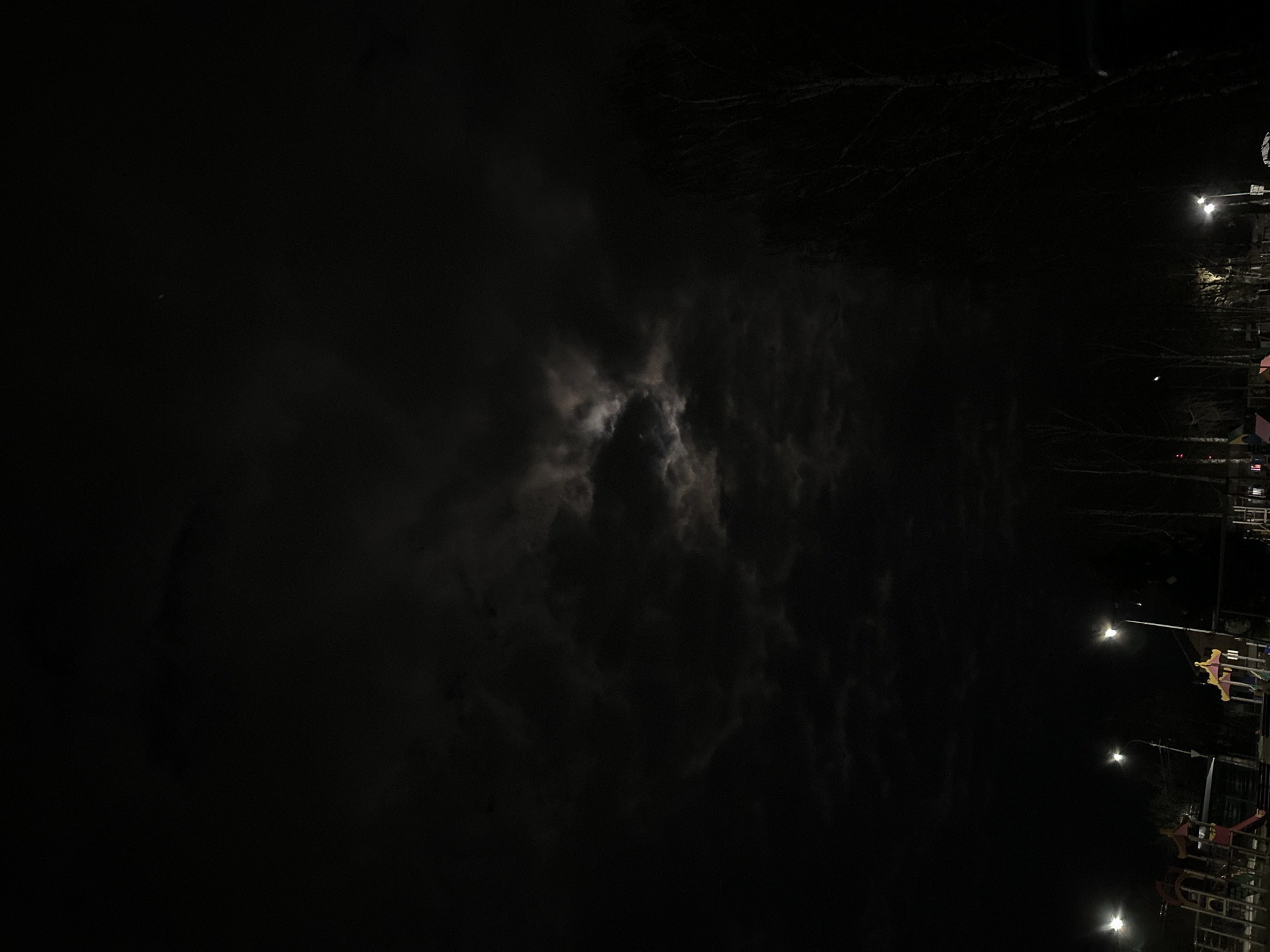 A night scene with a sky dominated by dark clouds partially obscuring the moon, with street lights and buildings visible on the right side. The location is likely Chaikovsky, Russia based on the prompt. The street lights illuminate a small area, suggesting a street or park area. The buildings are silhouetted against the night sky.