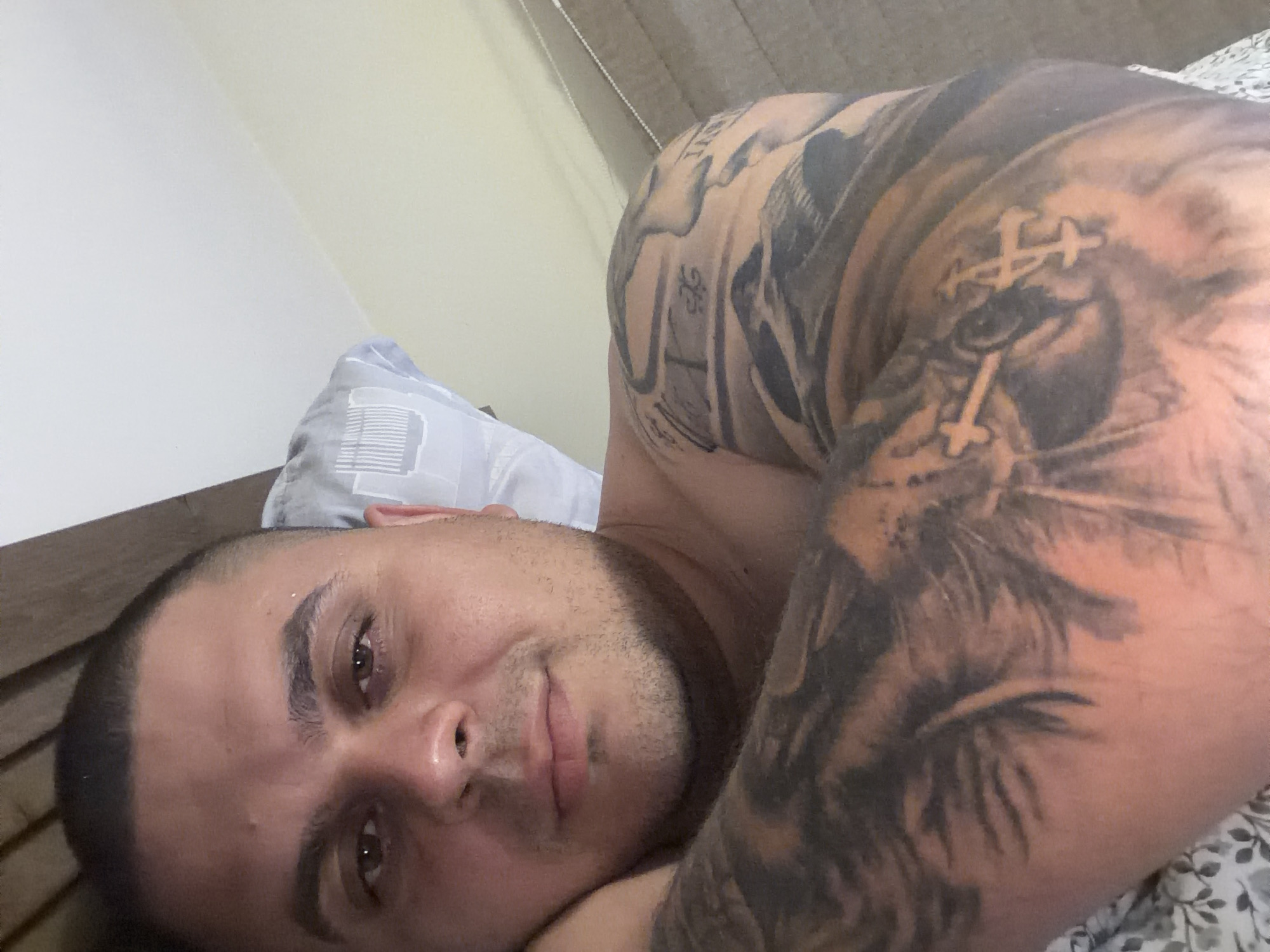 A man is captured in a close-up selfie, lying down in what appears to be a bedroom. He looks directly at the camera with a relaxed and slightly smiling expression. His head rests on a light-colored pillow featuring a subtle geometric pattern. A dark wooden bed frame or headboard is partially visible to his left.
The man has short, dark hair with a faded haircut on the sides, and light stubble covers his face. He is shirtless, prominently displaying a large, intricate black and grey tattoo that extends across his right arm and shoulder. The tattoo design includes what appears to be a lion's face or head, integrated with several cross-like symbols (possibly Orthodox crosses), and other decorative patterns.
The background shows a plain light-colored wall, with a patterned bedsheet (featuring a floral or leafy design) visible on the right. The soft, ambient lighting suggests it is daytime. The overall scene conveys a moment of calm and rest, likely taking place in a private residence in Shumen, Bulgaria. There is no discernible text visible in the image.