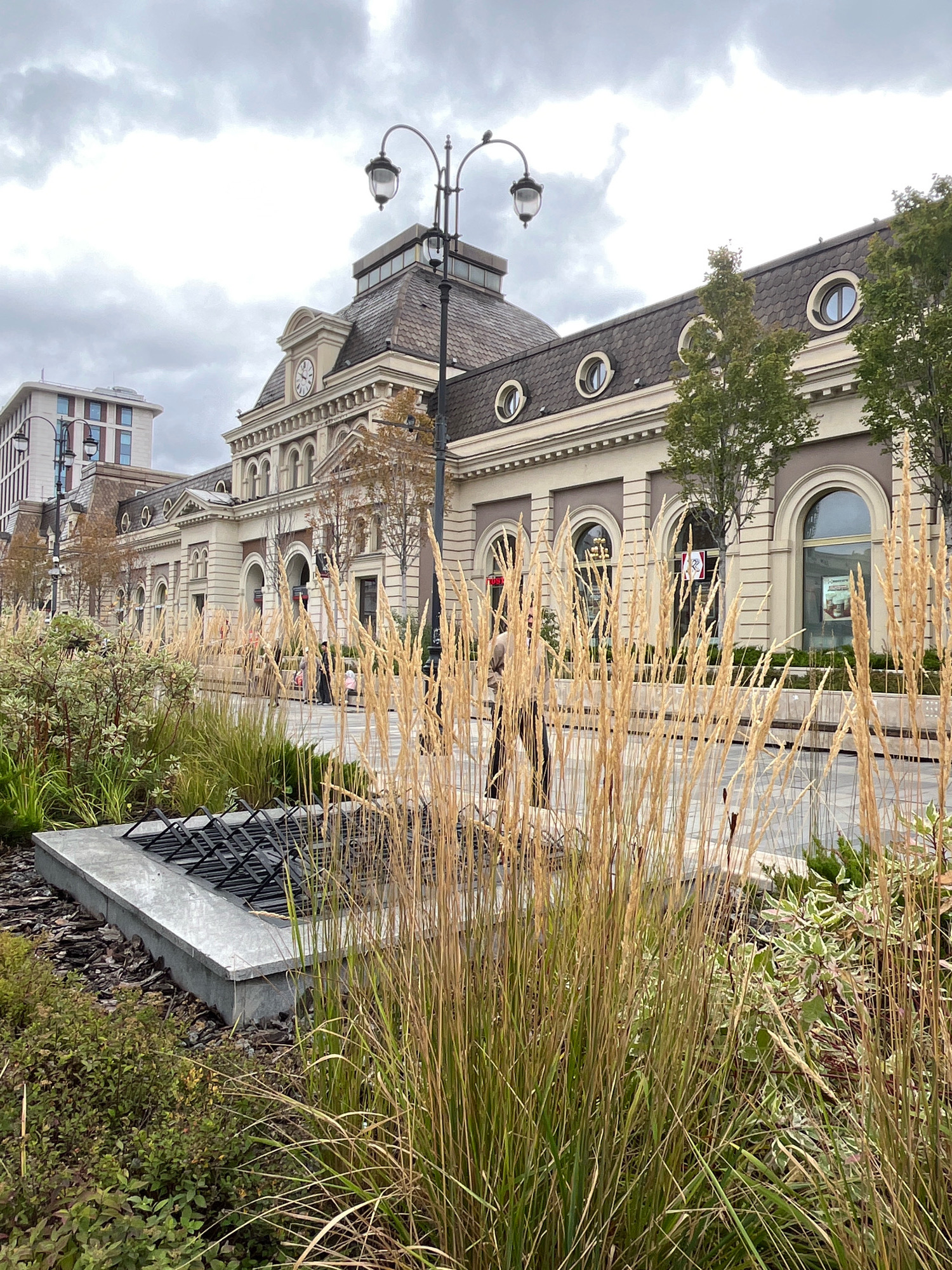 The image captures a view of the former VDNKh, now renamed Moscow City Business Center, in Moscow, Russia. The foreground is dominated by tall, dry grass, with a concrete planter containing a black metal structure, possibly for seating or decorative purposes, and surrounding mulch and greenery. Behind the grass, a paved walkway is visible, with a few scattered people walking. In the background, a grand, ornate building with a mansard roof, dormer windows, and a clock tower stands prominently. The building's facade features arched windows and decorative molding. To the left, a modern, multi-story building with a grid-like facade is visible. The sky is overcast with gray clouds.