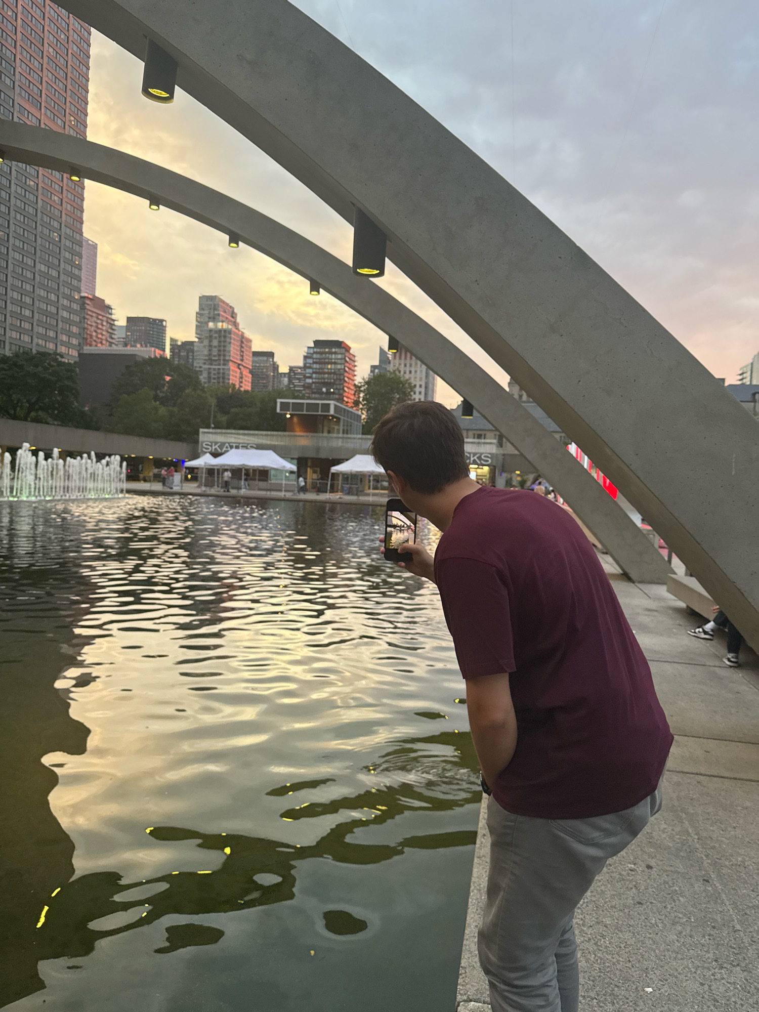 An outdoor urban scene in Toronto, Canada, captures the warm hues of sunset reflecting on a body of water. In the foreground, a person, viewed from behind, is leaning over the edge of the water, holding a smartphone to take a picture. They are wearing a maroon t-shirt and light grey pants. The water is calm with gentle ripples, and a fountain is visible in the background, to the left, with multiple jets of water reaching upwards. Beyond the fountain, a streetscape unfolds with several tall buildings and some tents. A concrete structure with a curved design arches over the scene. The sky is cloudy and tinged with orange and pink from the setting sun. The overall ambiance is peaceful, with a focus on the interplay of light and water.