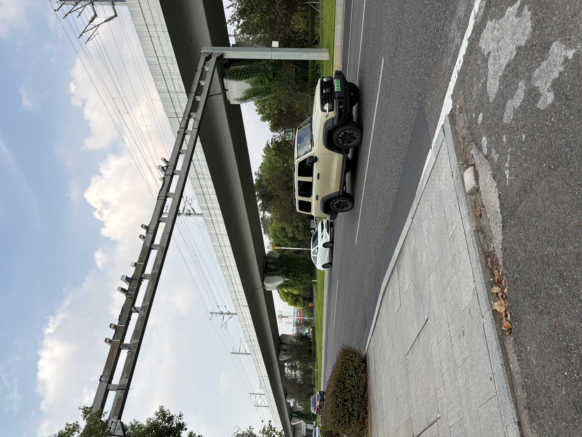 A light-colored SUV, identified by license plate "AA 667518", is traveling on a multi-lane paved road in Jiangjin, China, beneath an elevated railway line. The SUV is in the right lane, with other vehicles visible further down the road, including a red sedan and a white sedan. The road has white lane markings. To the left of the road is a paved sidewalk with granite tiles, bordered by a strip of darker asphalt and vegetation. Overhead, the elevated railway structure is supported by concrete pillars covered in green ivy. The sky above is partly cloudy.