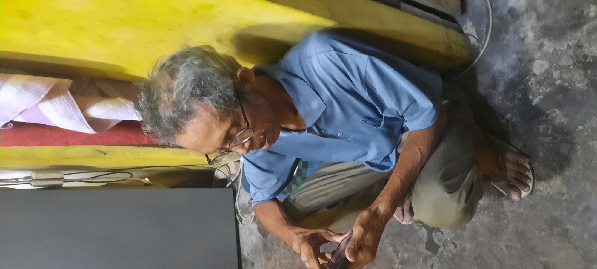 The image depicts an elderly man with graying hair, wearing glasses and a blue collared shirt, sitting on a rough concrete floor. He is holding a dark-colored smartphone in his hands and appears to be looking at it. The man is crouched down, with his legs bent and his feet, clad in light-colored sandals, visible on the floor. Behind him, a rough, textured yellow wall is prominent, with a section of a red wall and a striped towel hanging to the left. A grey object, possibly a piece of furniture or appliance, is visible to the far left. Electrical cords are present near the yellow wall. The overall environment suggests an interior space, likely a dwelling, with a utilitarian aesthetic. The scene is captured from a slightly elevated perspective, looking down at the man.