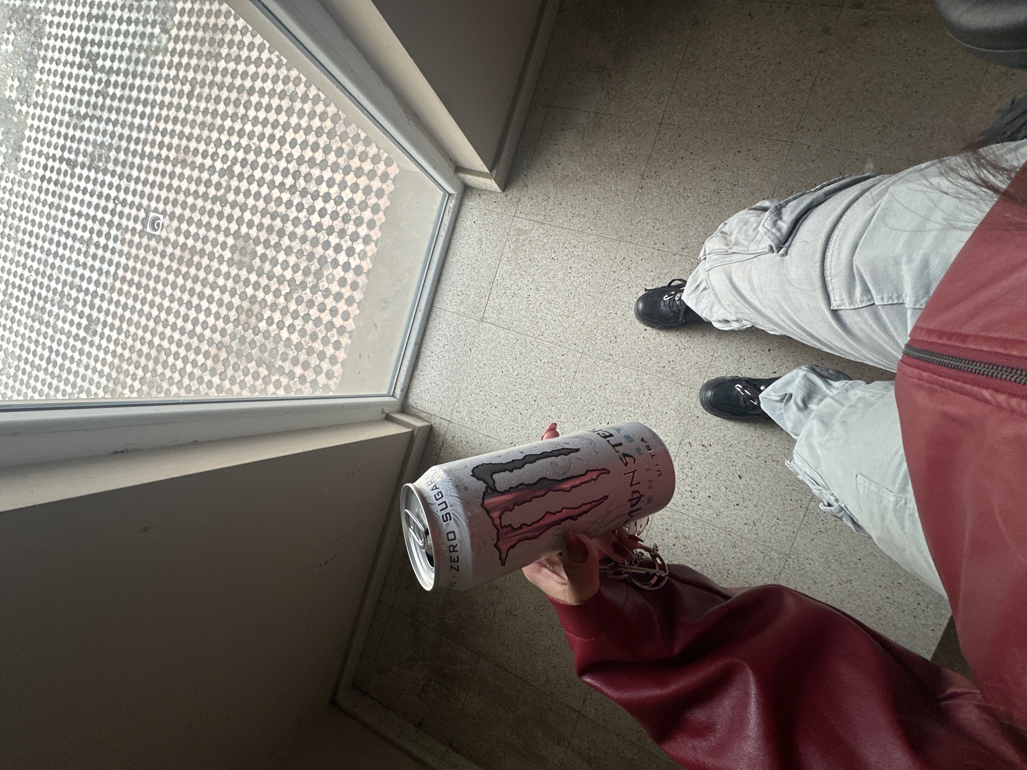 A person is standing inside a building near a window, holding a can of Monster Energy Ultra Zero Sugar drink. The person is wearing a red jacket and light grey cargo pants with black shoes. The floor is a light grey tile. The window has a pattern of small grey circles on it. The location appears to be indoors, possibly a hallway or entrance area. There is no information to confirm this is in Sofia, Bulgaria.