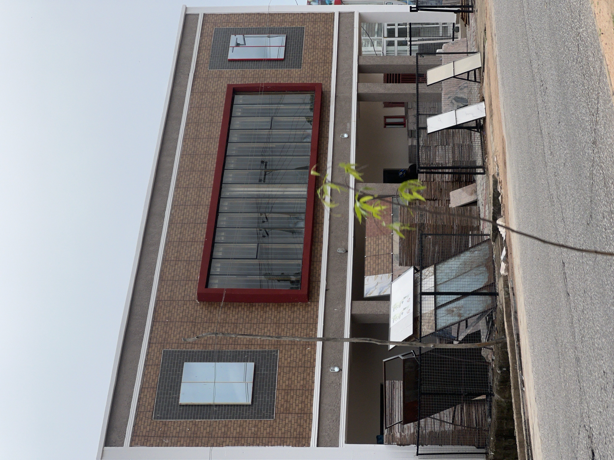This is a photograph of a building facade and foreground in Jalingo, Nigeria. The building is multi-story with a modern design, featuring a combination of brown brick-patterned siding and grey stucco. There are several windows visible; a large, rectangular window with vertical blinds is centrally located and framed in red. Another window, appearing to be a single pane, is positioned to the left, set within a dark grey textured panel. To the right, a smaller, vertically oriented window is also visible. The ground floor of the building appears to be an open-air space, with visible supports and some construction materials.
In the foreground, a black metal fence encloses an area containing stacks of construction materials, possibly tiles or planks, and some metal bins. There are also two large, rectangular panels, one white and one cream-colored, propped up on stands. A thin, bare tree branch extends into the frame from the lower right. The area in front of the fence is paved with a grey road and a concrete curb. The sky is overcast and pale. There are no discernible people actively engaged in activity. The overall impression is of a commercial or residential construction site, or a newly completed building with ongoing landscaping or finishing work.