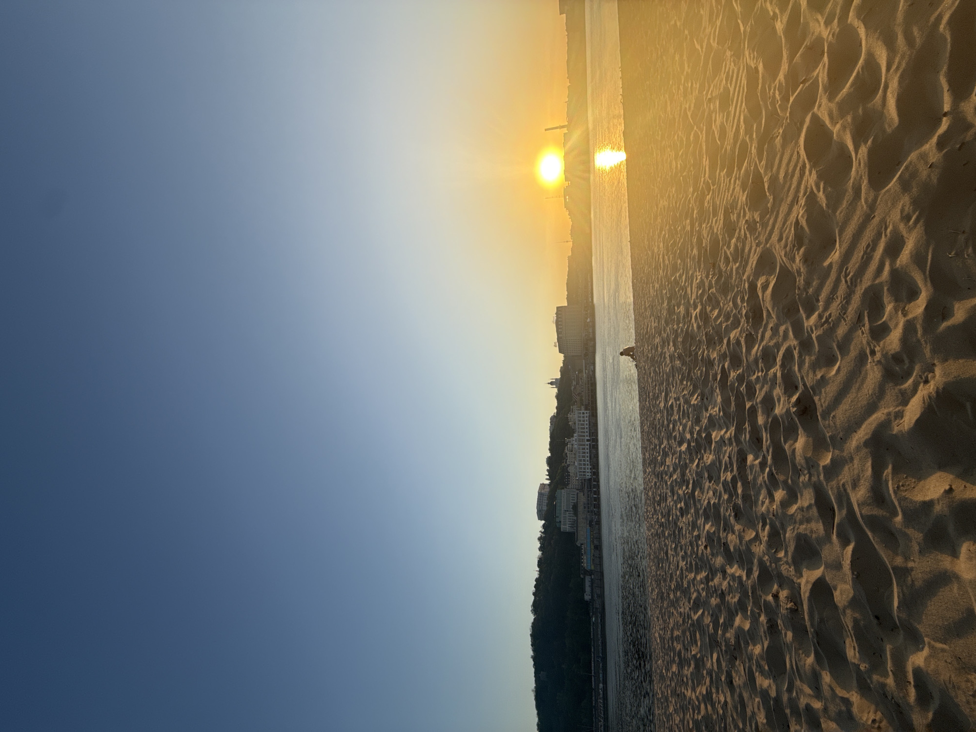 The image depicts a beach scene at sunset in Kyiv, Ukraine. The sun is low on the horizon, casting a golden light across the scene. The beach consists of sand with visible footprints and textures. In the distance, buildings and structures are silhouetted against the bright sun. The sky transitions from a dark blue to a lighter gradient near the horizon where the sun is positioned. A body of water reflects the sunlight. In the central area of the image, a bird is standing on the beach along the shoreline.