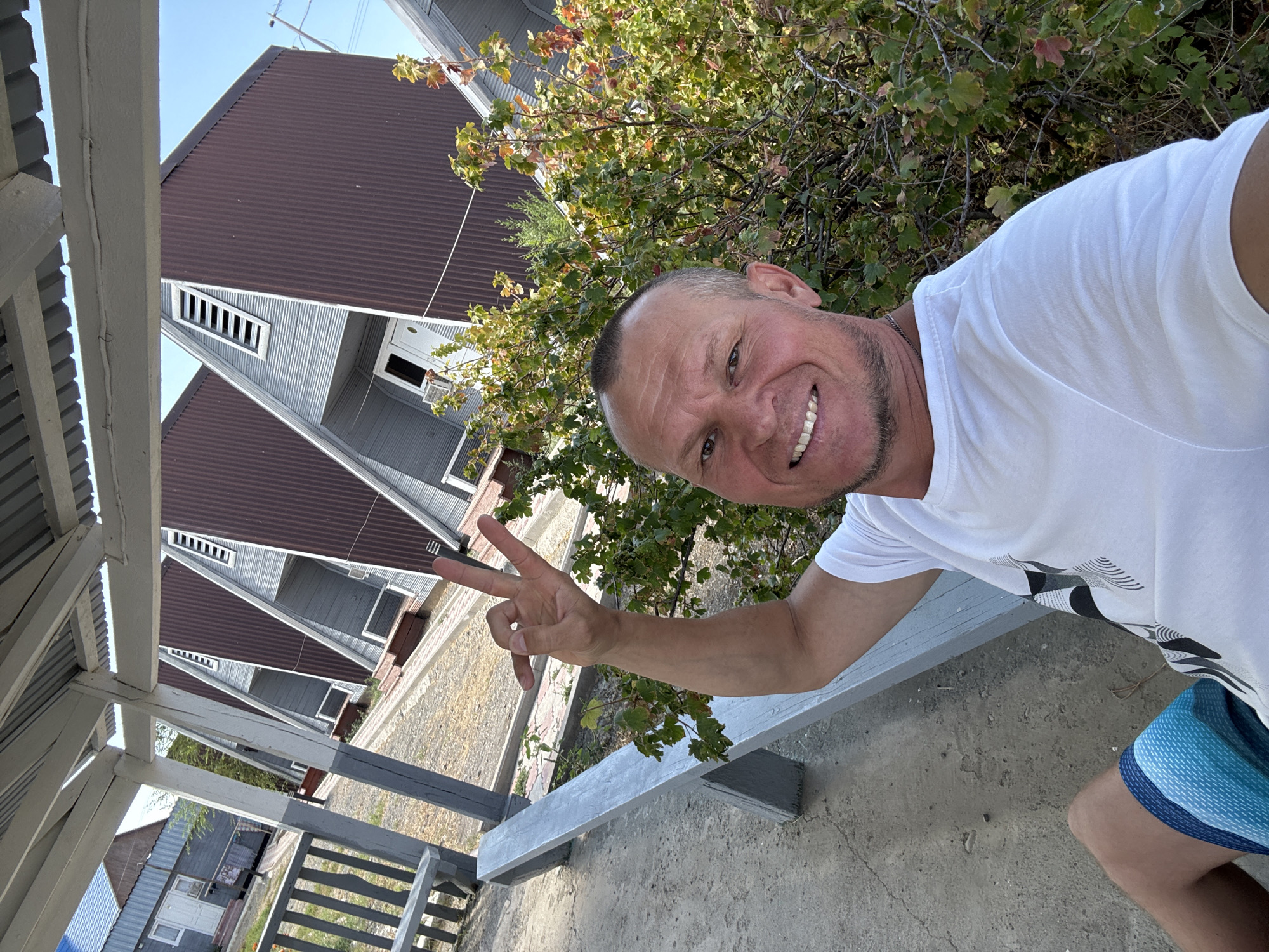A smiling man is taking a selfie outdoors. He is wearing a white t-shirt and blue, patterned swim trunks. His left arm is raised, and he is making a peace sign with his fingers. He is seated on a light-colored bench under a covered, open-air structure. In the background, several triangular-shaped buildings with brown corrugated metal siding and grey siding are visible. Green foliage is also present. The ground appears to be gravel and concrete. The sky is clear and blue. The scene suggests a vacation or resort setting.