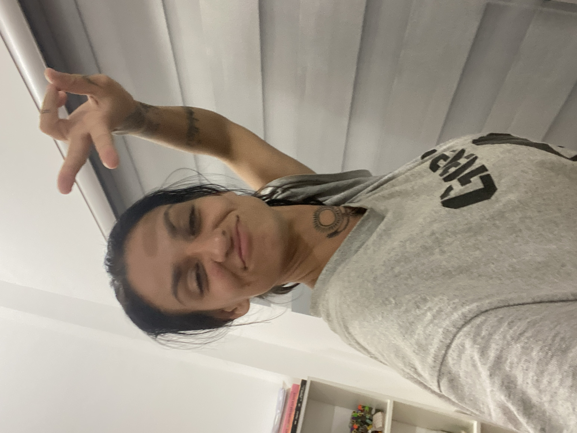 A woman with dark hair and a tattoo on her neck poses for a selfie. She is wearing a gray t-shirt and has her right arm raised, making a gesture with her fingers. The background appears to be an interior wall with horizontal blinds. The image seems to have been taken in an indoor setting, possibly a home or hotel room in Mũi Né, Vietnam, given the information provided. There is a white shelf with books and some decorative items visible on the left side of the frame.