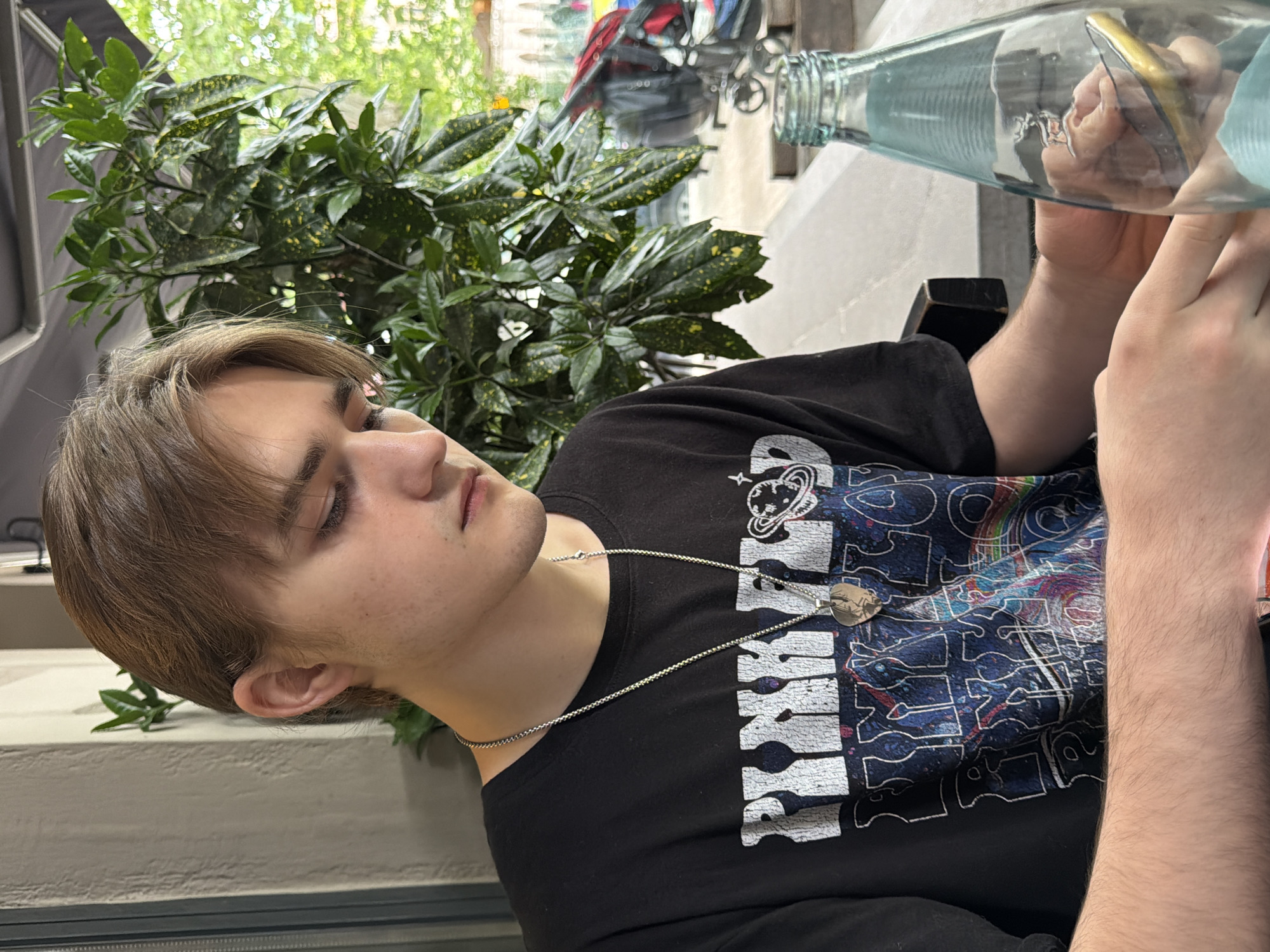 A young man sits at an outdoor table, likely at a café, in Tbilisi, Georgia. He is wearing a black t-shirt with a white graphic and a silver necklace. He is holding a glass bottle filled with water, in his hands to his side. A potted plant sits to his left. In the background, a street scene is visible, featuring pedestrians and vehicles.