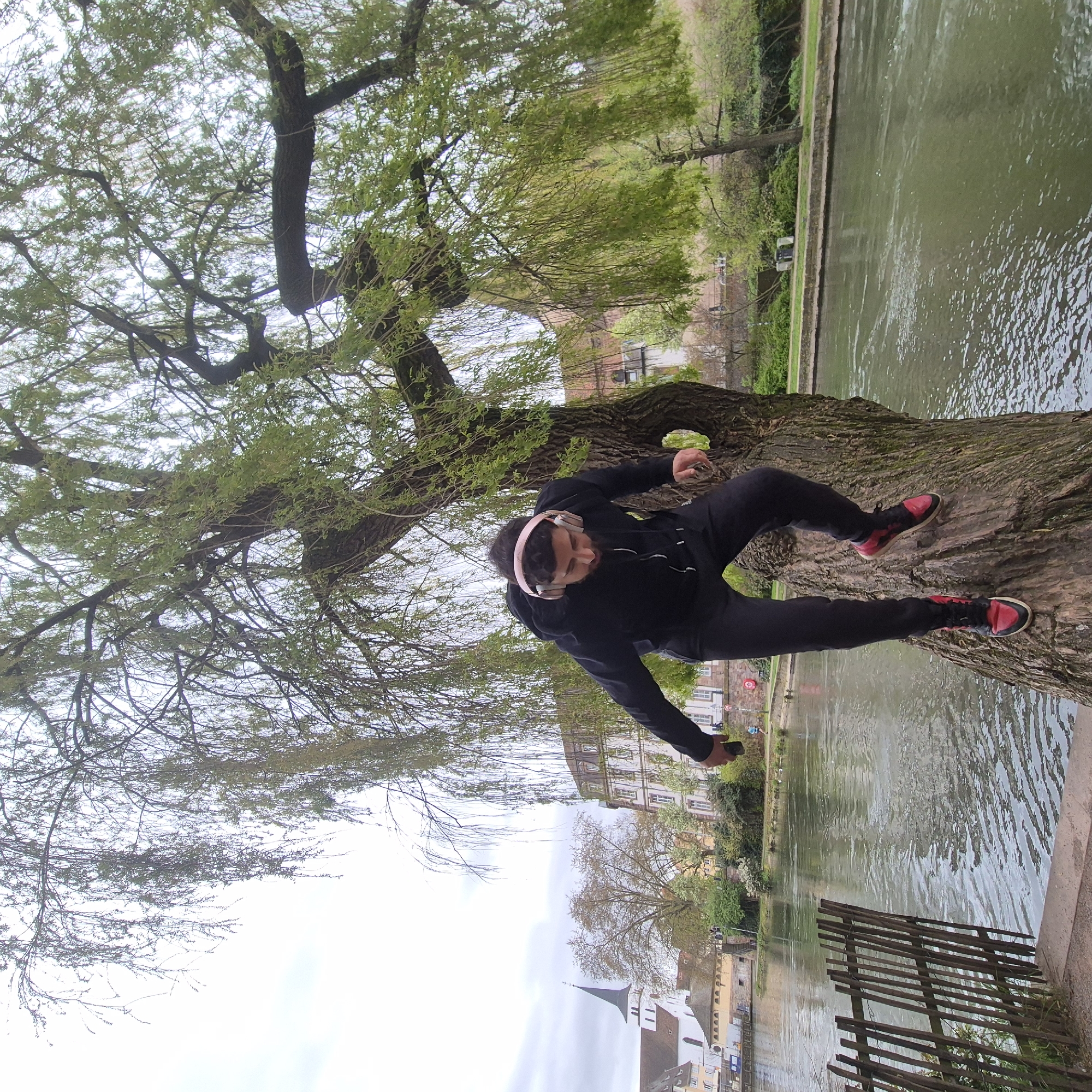 The image captures a person sitting on the trunk of a large tree, likely a weeping willow, which is positioned near a river or canal. The person, wearing a black jacket and pants, is also wearing pink headphones and red and black sneakers. They appear to be holding a smartphone.
The background features buildings typical of Strasbourg, France, including a distinctive church with a pointed spire. The river or canal is visible, and a rustic wooden fence or barrier runs along the bank in the foreground. The sky is overcast, suggesting a cloudy day.
