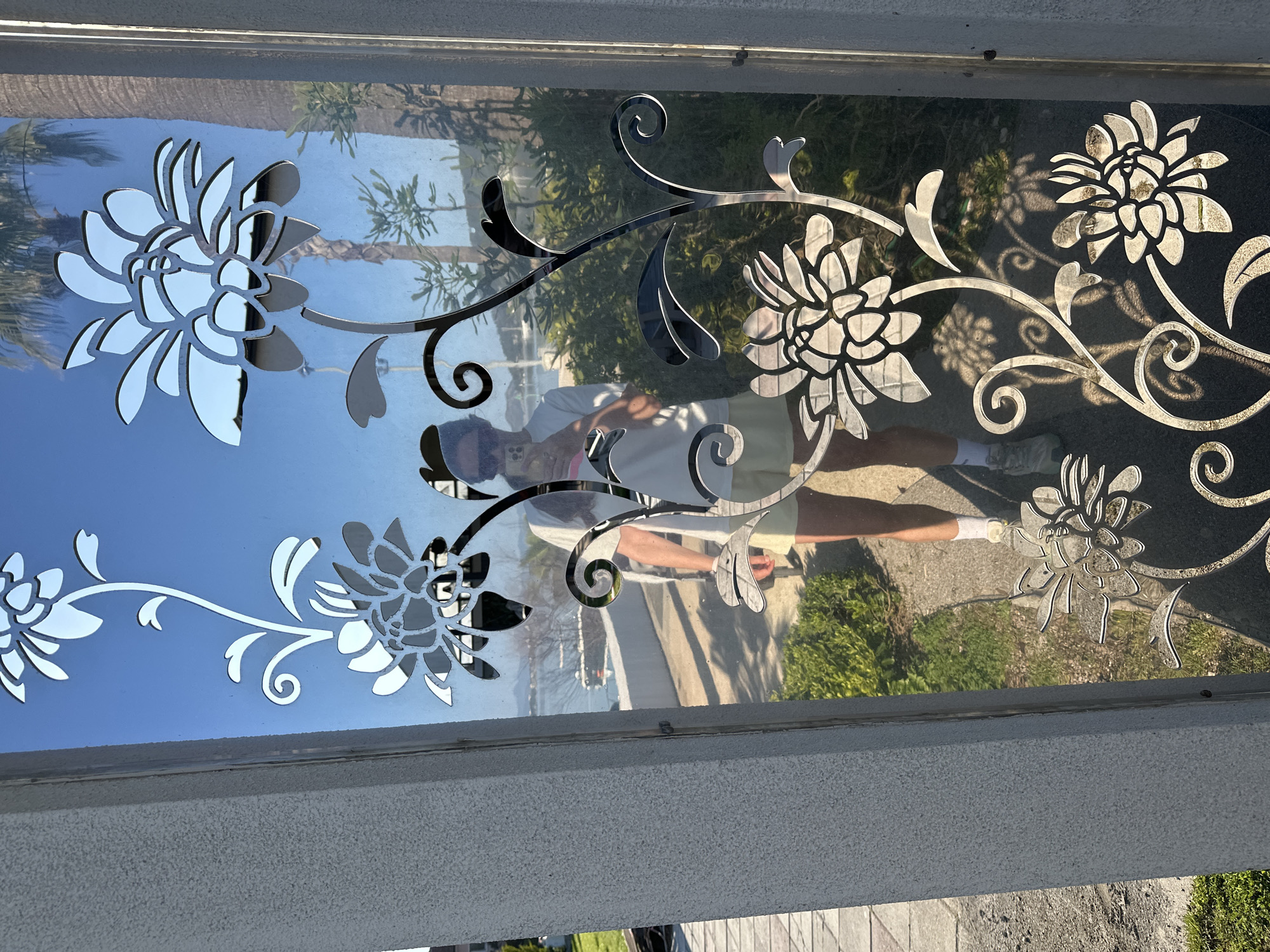 The image depicts a decorative glass panel with floral designs, likely part of a door or window. The panel has a metallic, reflective quality, showing a reflection of the person taking the photo and the surrounding environment. The person is wearing a light-colored shirt and shorts, white socks, and appears to be holding a mobile phone. The environment outside the glass panel includes foliage, trees, and a paved area. The foliage is characteristic of a warm, possibly Mediterranean climate. Given the prompt constraints, it is not possible to ascertain the specific location within the city of Fethiye, Turkey.
