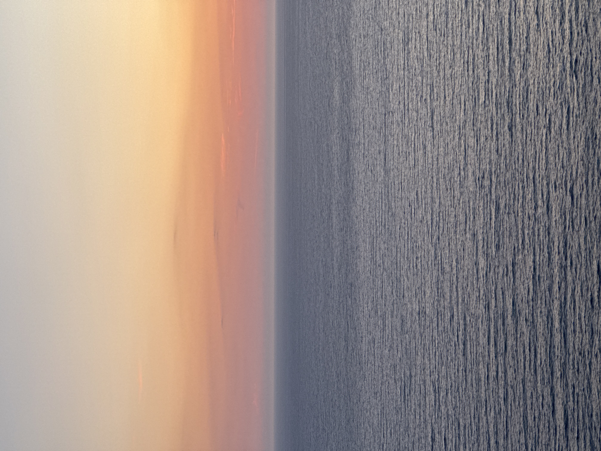 A serene seascape is depicted, with the ocean filling the lower two-thirds of the frame and the sky occupying the upper third. The ocean's surface is calm, exhibiting a gentle ripple pattern that stretches to the horizon. The water's color is a deep, muted blue-gray.
The sky above transitions from a soft, pale gray at the very top to a gradient of warm hues. The colors range from a pale yellow-orange near the horizon, blending into soft pinks and peachy tones, and finally fading into the pale gray. A few faint, wispy clouds are present, subtly catching the light.
The image captures the tranquil atmosphere of a coastal scene, likely at dawn or dusk, in the vicinity of Glavatičići, Montenegro. There are no people, specific objects, or actions visible in the frame; the focus is solely on the natural expanse of the sea and sky.