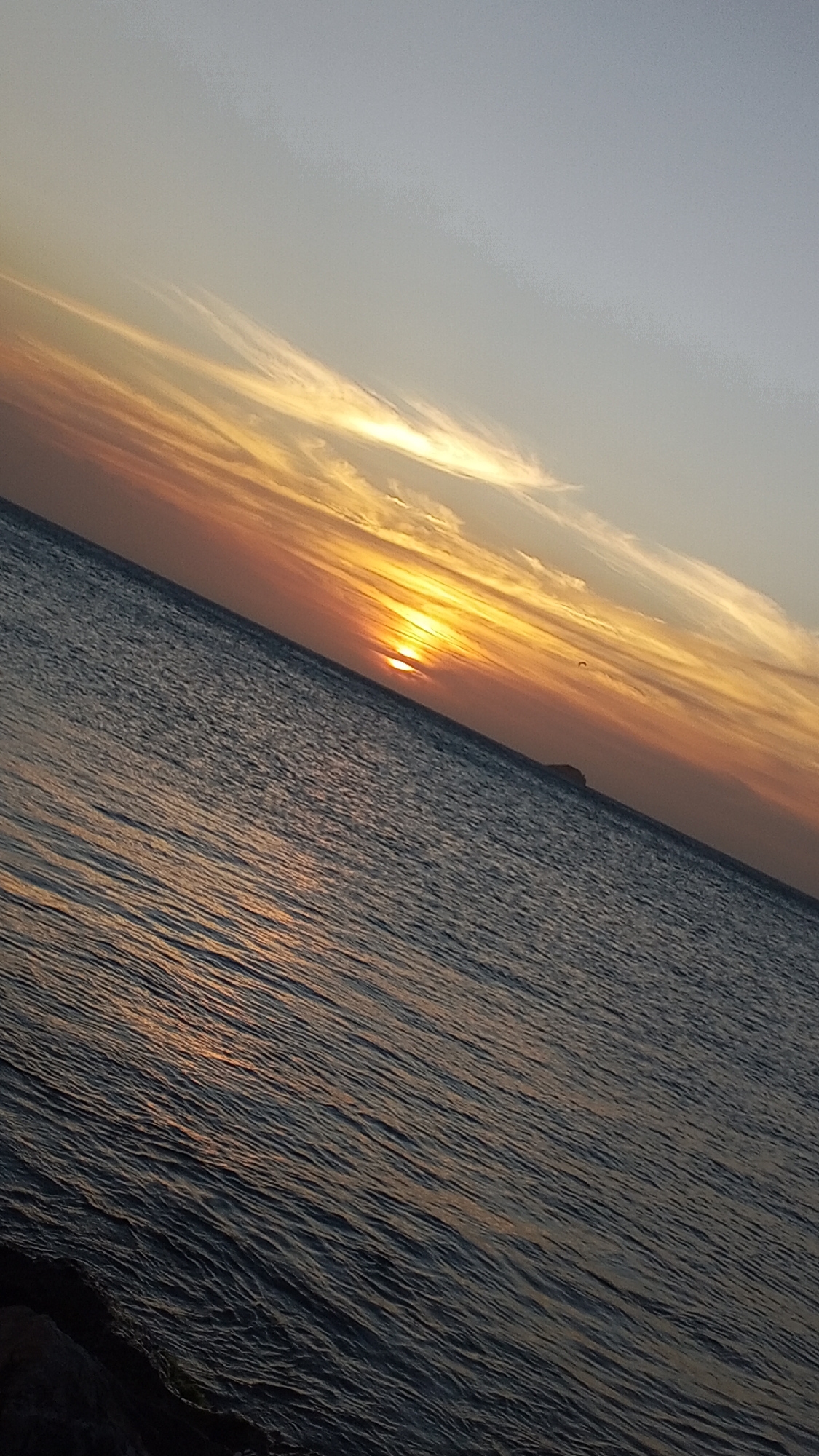 The image depicts a sunset over the ocean. The sun is low on the horizon, partially obscured by a cloud layer, creating an orange and yellow glow. The sky ranges from light gray to orange. The ocean reflects the colors of the sunset. There is a small landmass visible on the horizon. Rocks are in the lower left corner. The location is likely Pedro González, Venezuela.