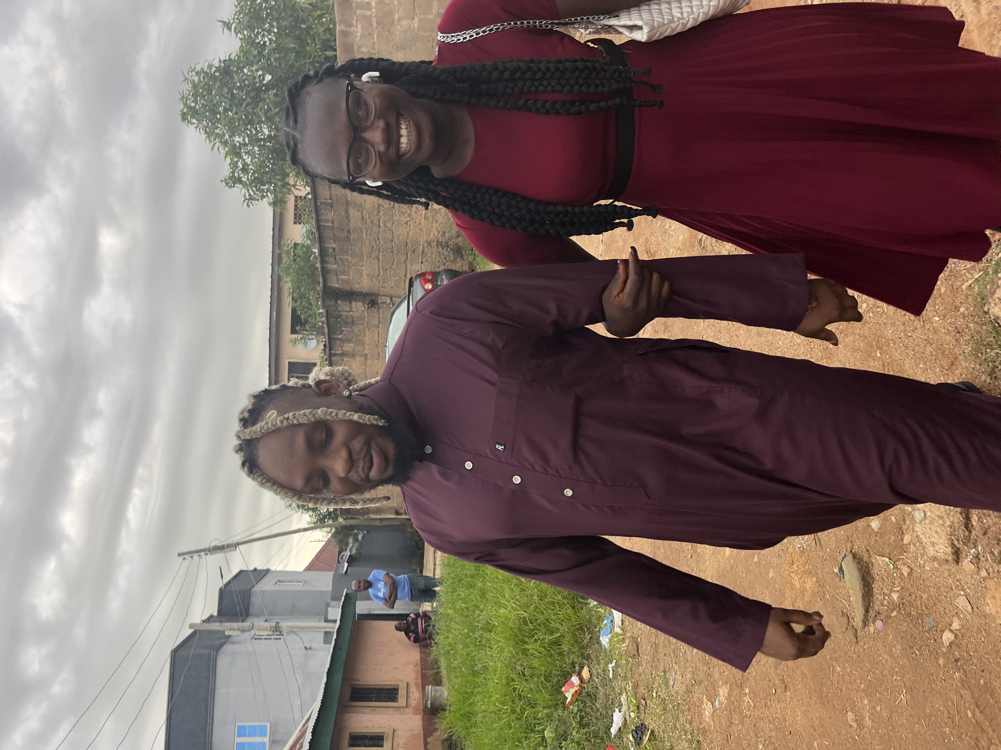 A medium shot captures a Black man and a Black woman walking together outdoors on a dirt path in Jos, Nigeria. The man, on the left, wears a long-sleeved, burgundy traditional outfit. He has light-colored dreadlocks and is looking down and to his left. The woman, on the right, wears a burgundy dress and glasses. She has long, dark braided hair and is smiling at the camera. She carries a white purse on her right shoulder. The man has his left arm linked with the woman's right arm. In the background, there are residential buildings, a brick wall, a car, and some overgrown vegetation. The sky is overcast.