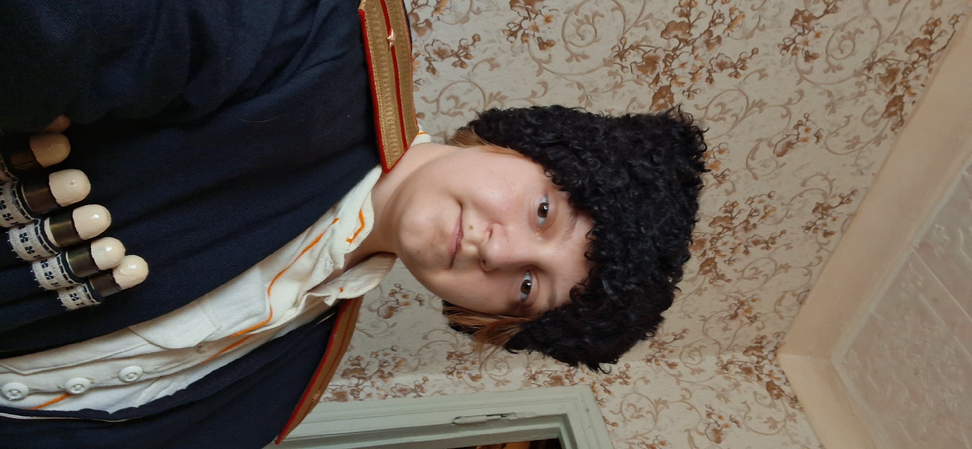 A person is wearing a black fur hat, a white shirt with orange trim, and a dark blue jacket with gold and red epaulets. The person has light skin and shoulder-length reddish-brown hair. Several bullet-like objects are visible on a band across the jacket. The background is a wall with a floral wallpaper pattern. The image appears to be taken indoors, possibly in a residential setting.