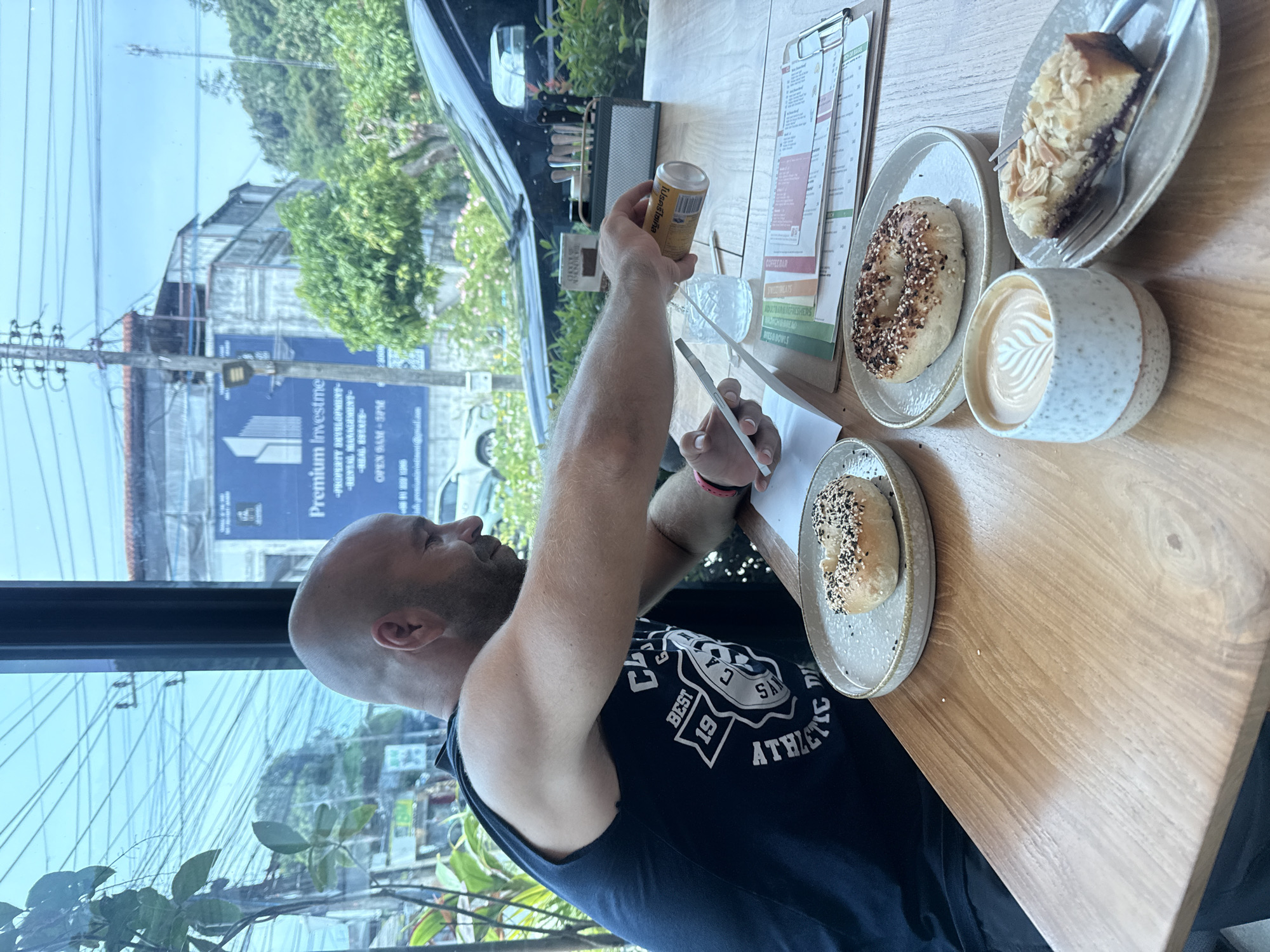 A bald man is seated at a wooden table in a cafe in Ko Samui District, Thailand, with a view of the street through a large window. He is wearing a dark blue tank top and is looking down at his phone, which he holds in his left hand. His right hand is holding a can and is in the process of pouring liquid into a glass. On the table in front of him are two bagels on separate plates, a slice of almond cake on a plate with a fork, and a cup of coffee with latte art. A menu is clipped to a board on the table. Outside the window, there are power lines, buildings, trees, and a black car.