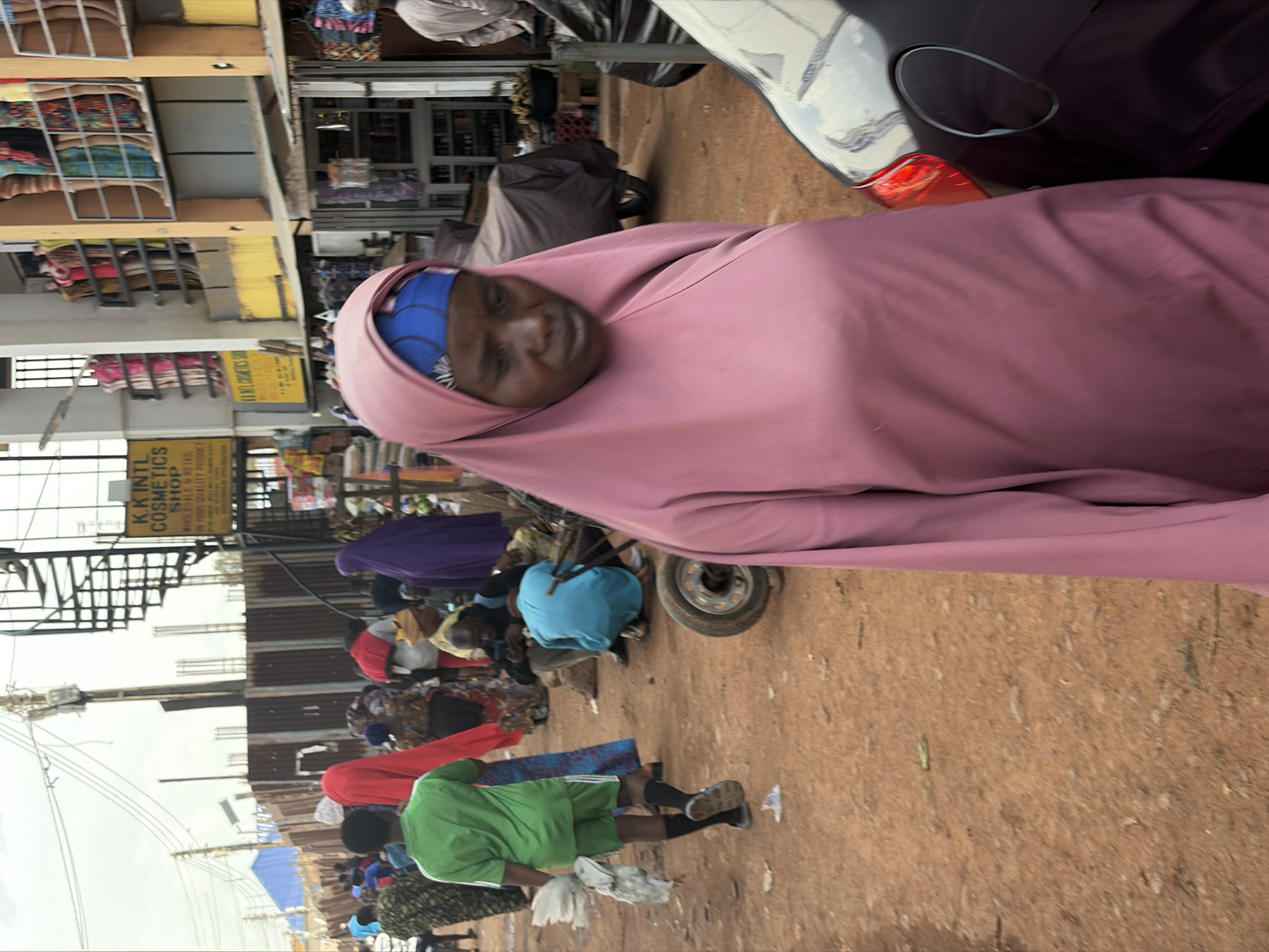 The image depicts a bustling outdoor market scene. The primary subjects are people dressed in various colorful clothing, many wearing headscarves or hijabs. In the foreground, a person is partially visible, draped in a large, loose pink garment, with a blue headscarf peeking out. Their face is turned towards the camera, and their expression appears neutral.
Behind this figure, other individuals are seen amidst stalls or makeshift selling areas. Some are gathered around, possibly browsing or interacting with vendors. One person is wearing a bright green shirt and shorts, and appears to be holding a white chicken. Another individual is dressed in a vibrant purple garment. The ground appears to be dirt or packed earth, characteristic of an outdoor market.
In the background, there are structures that suggest shops or market stalls, some with shelves displaying various items, possibly fabrics or merchandise. A vehicle, appearing to be a car or van, is parked on the right side of the image, with its rear visible. A yellow sign with text is attached to one of the stalls, reading "K.K. INTL COSMETICS SHOP. WHOLESALE & RETAIL. ON HIGH QUALITY PRODUCTS." The lighting suggests it is daytime, and the overcast sky indicates a potentially cloudy or humid weather. The overall atmosphere is one of everyday commerce and activity in a lively marketplace.
