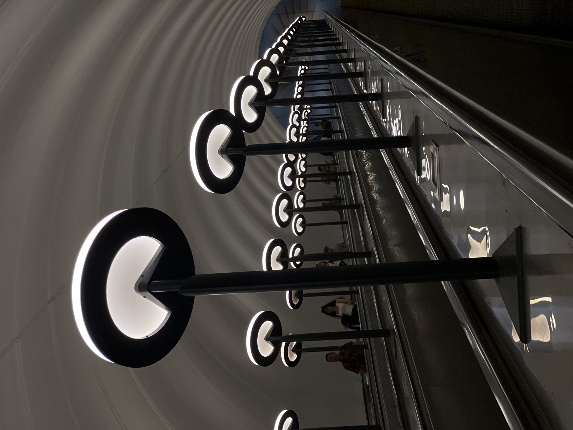 The image captures an escalator in a Moscow, Russia metro station. A line of Pac-Man shaped lights illuminate the ceiling on the left side of the escalator. The ceiling has a ribbed, curved design. On the right is the metal and glass side of the escalator. There are people riding on the escalator.