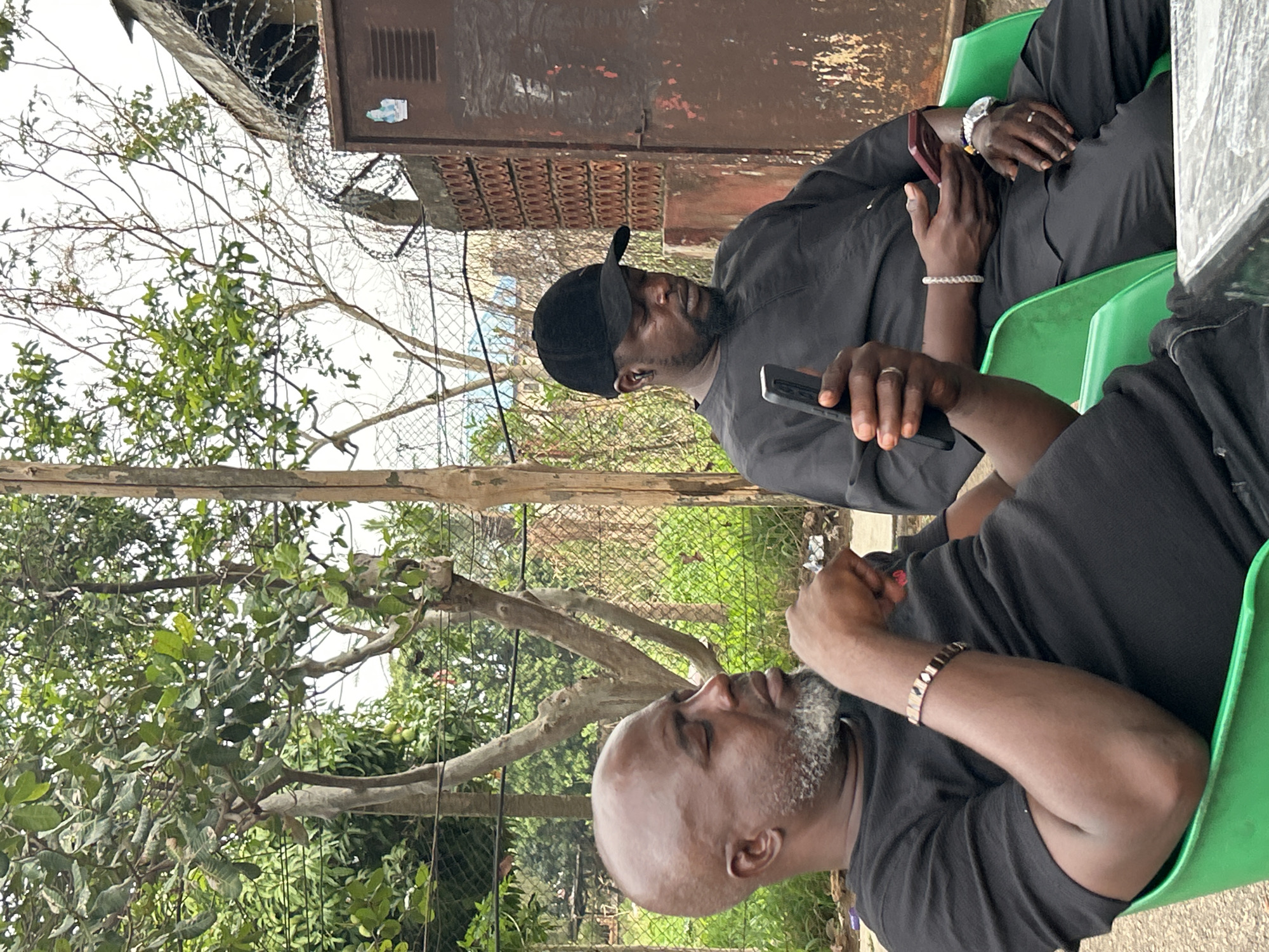 Two men sit on green plastic chairs outdoors in Jos, Nigeria. The man in the foreground, wearing a black T-shirt, has a shaved head and a gray beard. He is looking up and holding a black smartphone in his right hand. The second man, sitting behind him, is wearing a black baseball cap and a black long-sleeved shirt. He is also holding a black smartphone. Both men wear rings on their left hands and bracelets on their wrists. They sit beneath trees with green leaves. A chain-link fence and barbed wire top a concrete and corrugated metal structure.