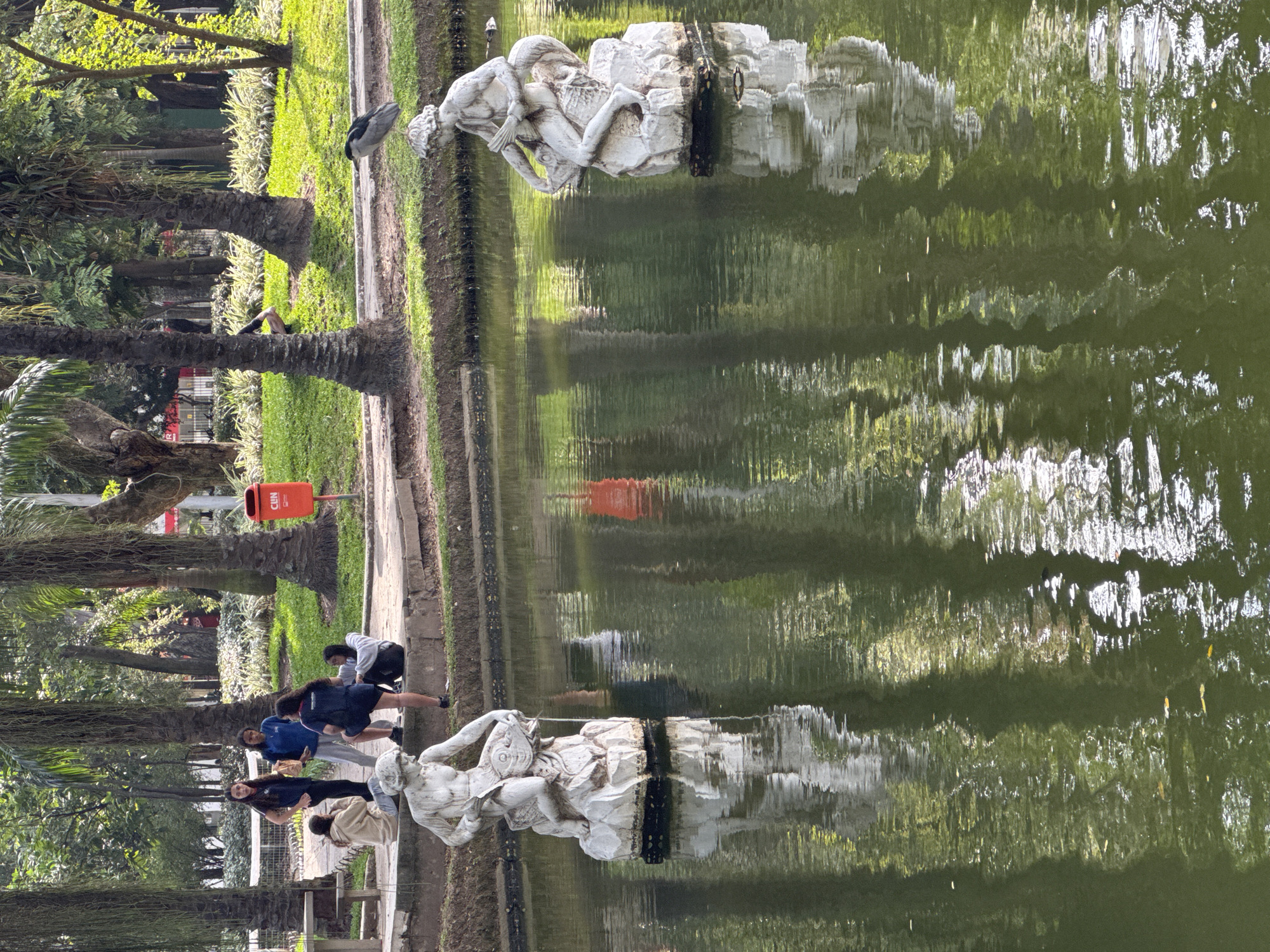 This is a medium-shot, eye-level, outdoor photograph of a park scene in Niterói, Brazil. In the foreground, two white stone statues are partially submerged in a green pond. The statue on the left depicts a nude male figure holding a fish, while the statue on the right shows a nude male figure seated on a rock with a fish. The water reflects the surrounding trees and sky.
In the midground, several people are gathered on a stone embankment. On the left, three young women stand together, one in a blue top and the others in dark tops. To their right, two young women are sitting on the embankment, one in a beige sweatshirt and the other in a dark top. Further to the right, a woman in a dark top and shorts stands with her back to the camera. Behind them, a red trash can with the word "CLIN" is visible next to a palm tree.
The background consists of lush greenery with numerous palm trees and other foliage. Sunlight filters through the trees, creating dappled light on the grass and ground. A white metal fence is visible behind the people on the embankment. The overall scene suggests a relaxed atmosphere in a public park.