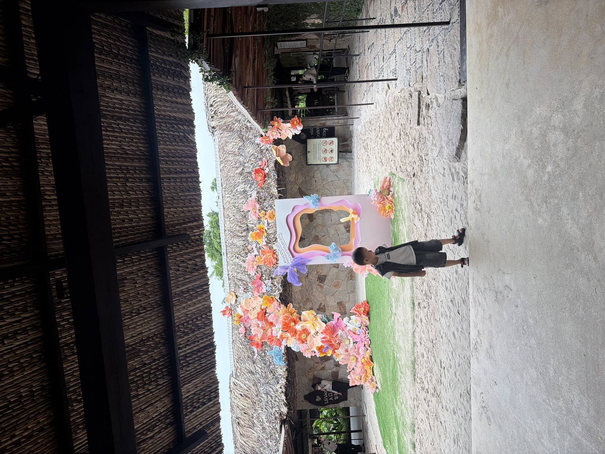 The image depicts a young boy, wearing a dark vest over a white t-shirt and grey shorts, standing on a sandy path. He is looking down and to his right. Behind him, there is an elaborate floral decoration of colorful paper flowers cascading down a thatched-roof building. A photo booth with a pastel frame is set up near the flowers. To the left, two people in uniform are standing near a stone wall with signage. The environment suggests an outdoor venue, possibly a resort or attraction, with a thatched-roof structure, stone walls, and a sandy ground. Based on the hashtag "#resortnhatrang" visible on the photo booth, the location is identified as Nha Trang, Vietnam.