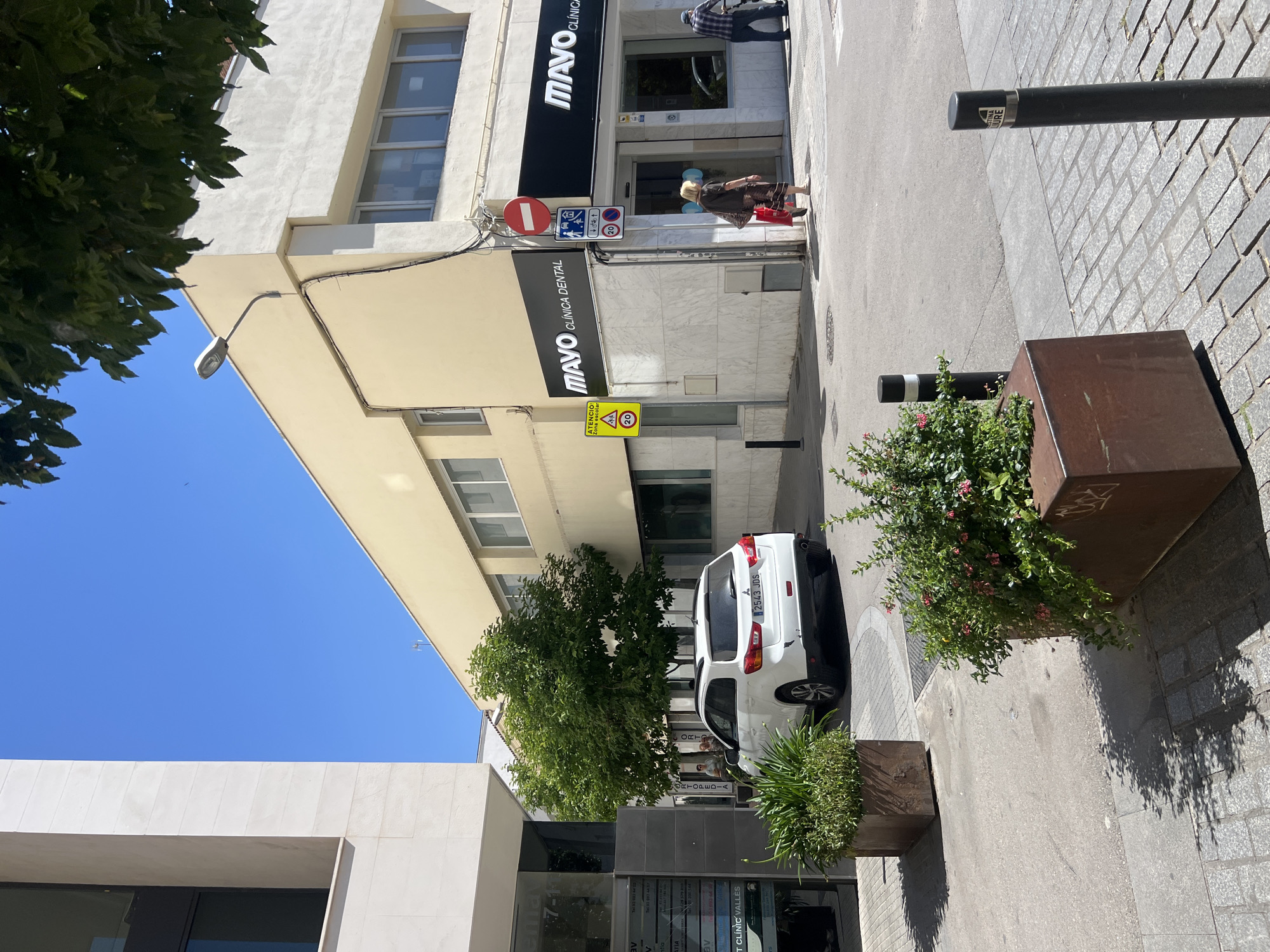The image depicts a street view in Granollers, Spain, specifically in front of the "MAYO CLÍNICA DENTAL" building. The building is multi-storied and cream-colored, and has a second sign that reads "MAVO CLÍNICA". A street lamp is affixed to the building's facade. Street signs indicating "Zona escolar" (school zone) and speed limits are posted near the clinic. A white car is parked along the curb. A few people are walking on the sidewalk near the clinic's entrance. There are bollards and planters with greenery are lining the sidewalk. The sky is clear blue. The architecture is modern.