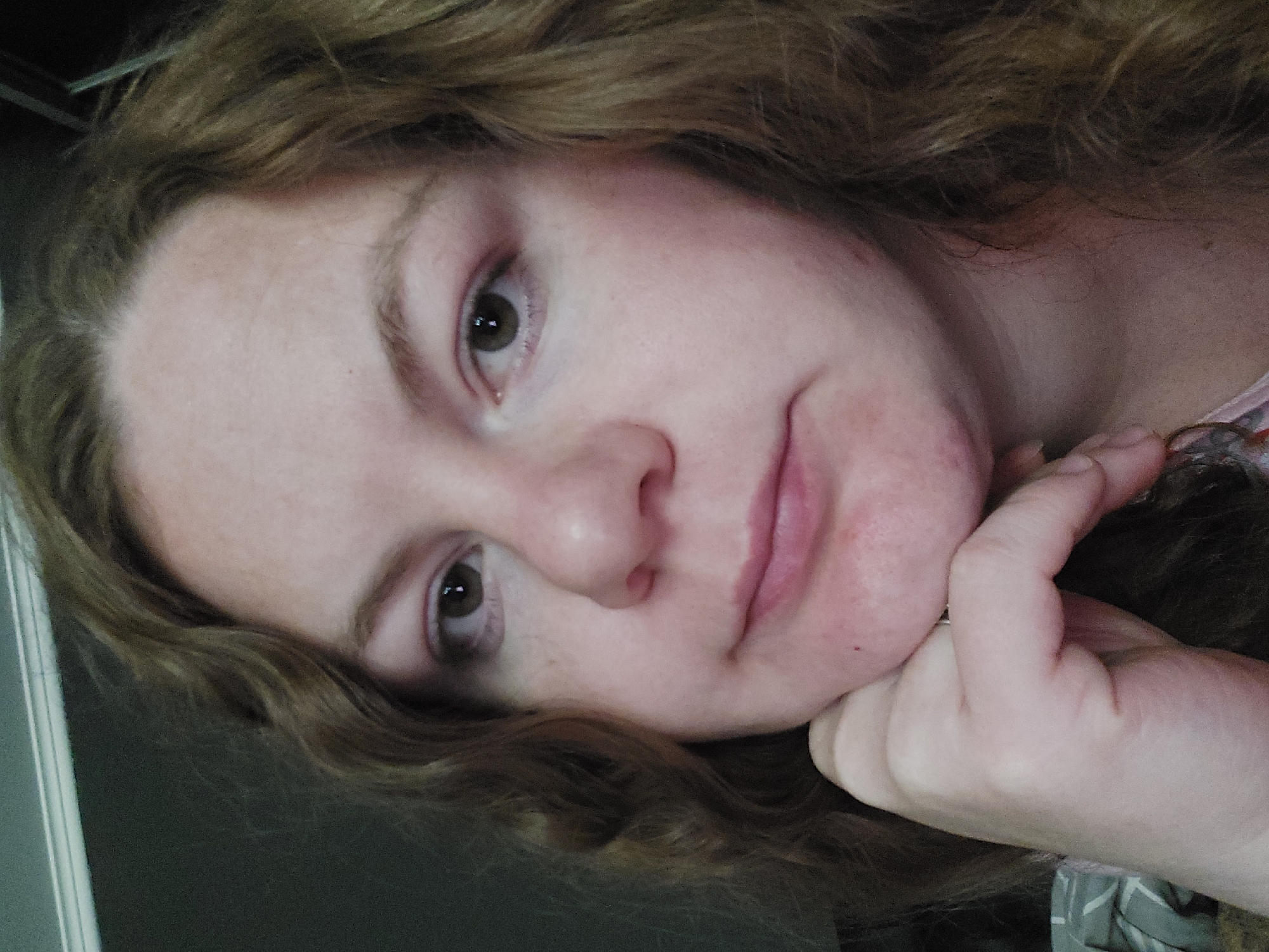 A medium close-up shot focuses on the face of a woman with wavy, light brown hair. She has fair skin with a rosy undertone, light brown eyes, and a small nose. Her hand is resting under her chin, with her fingers slightly curled. Her lips are closed and appear to be lightly colored. The background is blurred, showing a dark wall with a white molding strip across the top. The lighting suggests the photo was taken indoors.