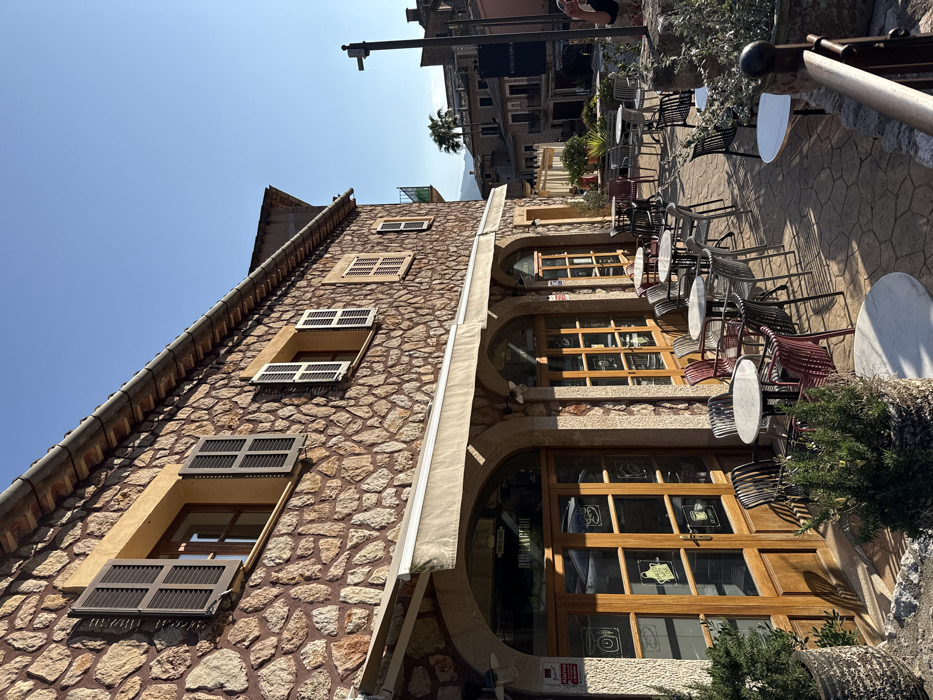 This image depicts a sunny outdoor scene in Fornalutx, Spain, focusing on a two-story building with a reddish-brown, irregular stone facade. The ground floor functions as a boulangerie or cafe, featuring three arched wooden storefronts with glass panes. White canvas awnings extend above these, providing shade. The leftmost storefront consists of double wooden doors with glass panes, adorned with white chalk-like drawings of a jar, bread, and a cup, with "BOULANGERIE" visible above. An alarm box is mounted to the wall left of the doors. The upper floor has multiple rectangular windows with light ochre surrounds, wooden frames, and dark grey louvered shutters; some are open, others partially closed. A white gutter traces the edge of the tiled roof.
A patterned light-brown paved area in front of the building accommodates an outdoor seating area. Numerous round tables with light-colored tops and dark bases are arranged with dark metal or plastic chairs in black, grey, and maroon. Several potted plants and shrubs are strategically placed around the seating and along a low stone wall to the right. In the background, a street leads to more traditional stone buildings, with a single palm tree visible among them under a clear, bright blue sky. A dark utility pole with an attached fixture stands on the far right. A dark sign with white lettering is partially visible on a distant building. One individual is partially discernible on the far right, their torso and arm (wearing a light-colored top) facing away from the viewer. Long shadows across the paving indicate either morning or late afternoon sun.