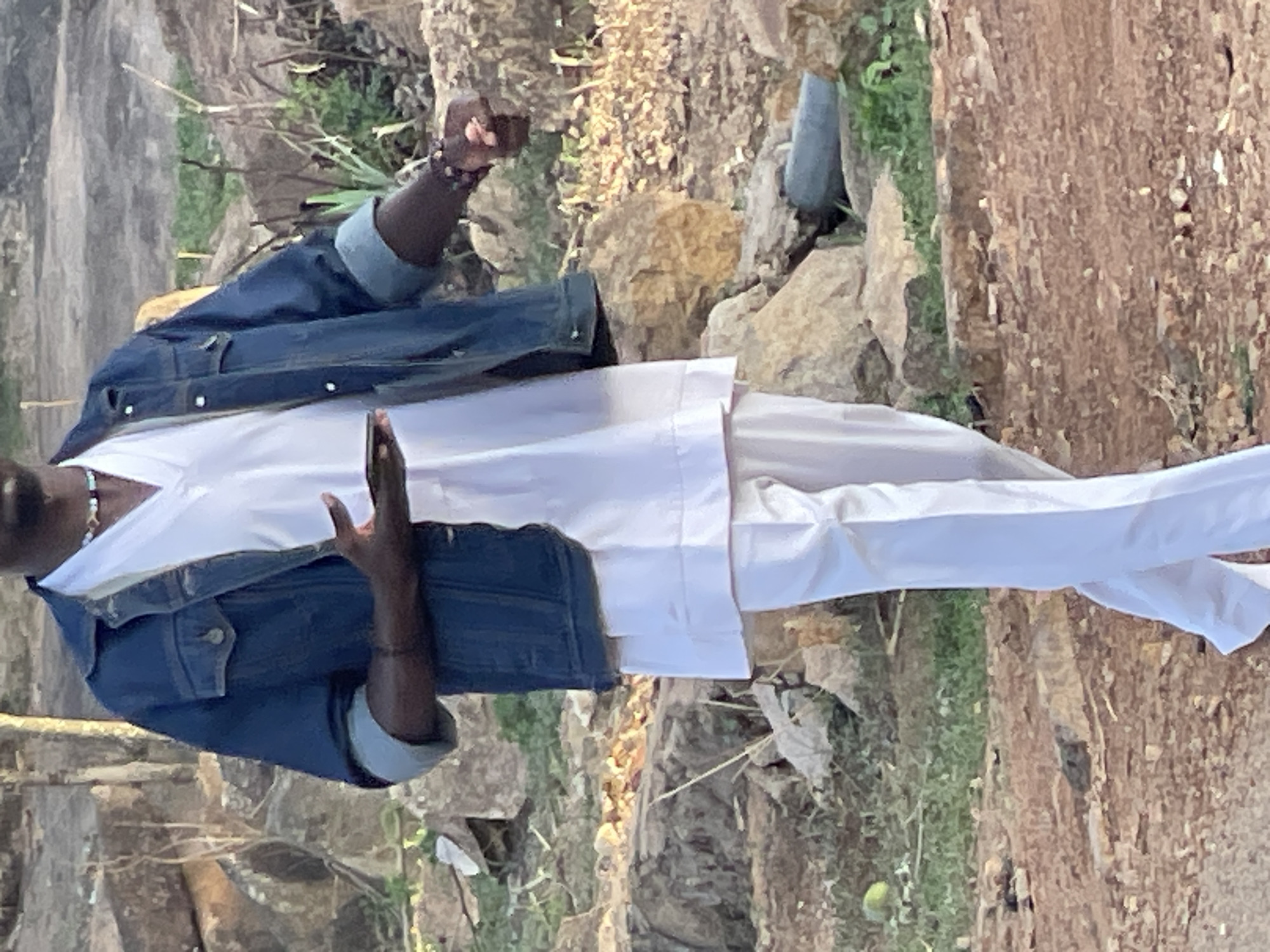 An African man wearing a blue jean jacket and white shirt and pants is standing outdoors. The background appears to be rocky terrain with green patches of vegetation. The location is likely near Jos, Nigeria, judging by the attire and terrain. The man's left hand is clenched into a fist while his right hand is extended in front of him with his palm facing towards the camera.