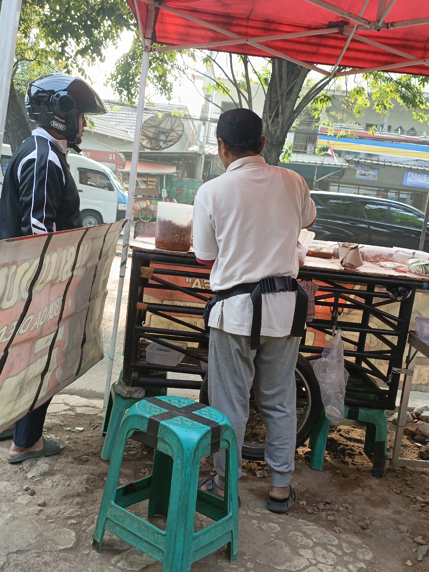 The image shows two individuals at a street food stall in East Jakarta, Indonesia. On the left, a person wearing a black and white jacket and a black helmet is standing. On the right, a person wearing a white t-shirt, gray sweatpants, and a black utility belt is attending to the food stall. The stall has a red canopy and displays various food items in containers, along with a paper-wrapped package. A green plastic stool is placed in front of the stall. The ground is paved with rough stones, and in the background, buildings, trees, and a passing white van are visible. The scene appears to be during daylight hours.