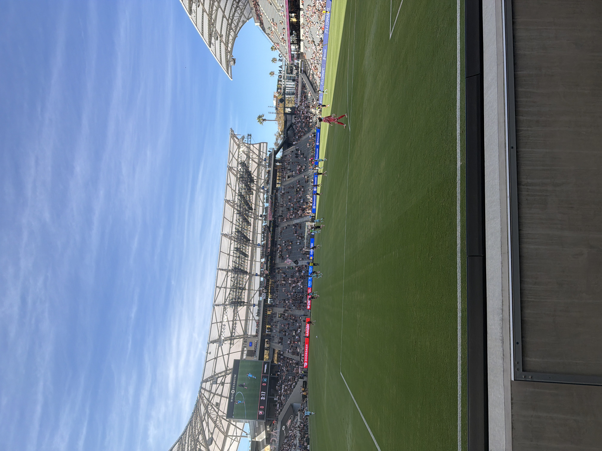 This is a photograph taken from the stands of BMO Stadium in Los Angeles, California, during a soccer match. The image captures a wide view of the green soccer field, with a large digital scoreboard displaying the game's progress in the background. Spectators are seated in the tiered stands surrounding the field, some of whom are wearing team colors. The stadium's modern architecture, including its curved roof and lighting structures, is visible against a bright blue sky with scattered clouds. A few palm trees are also visible beyond the stadium structure. The game appears to be in progress, with players visible on the field.