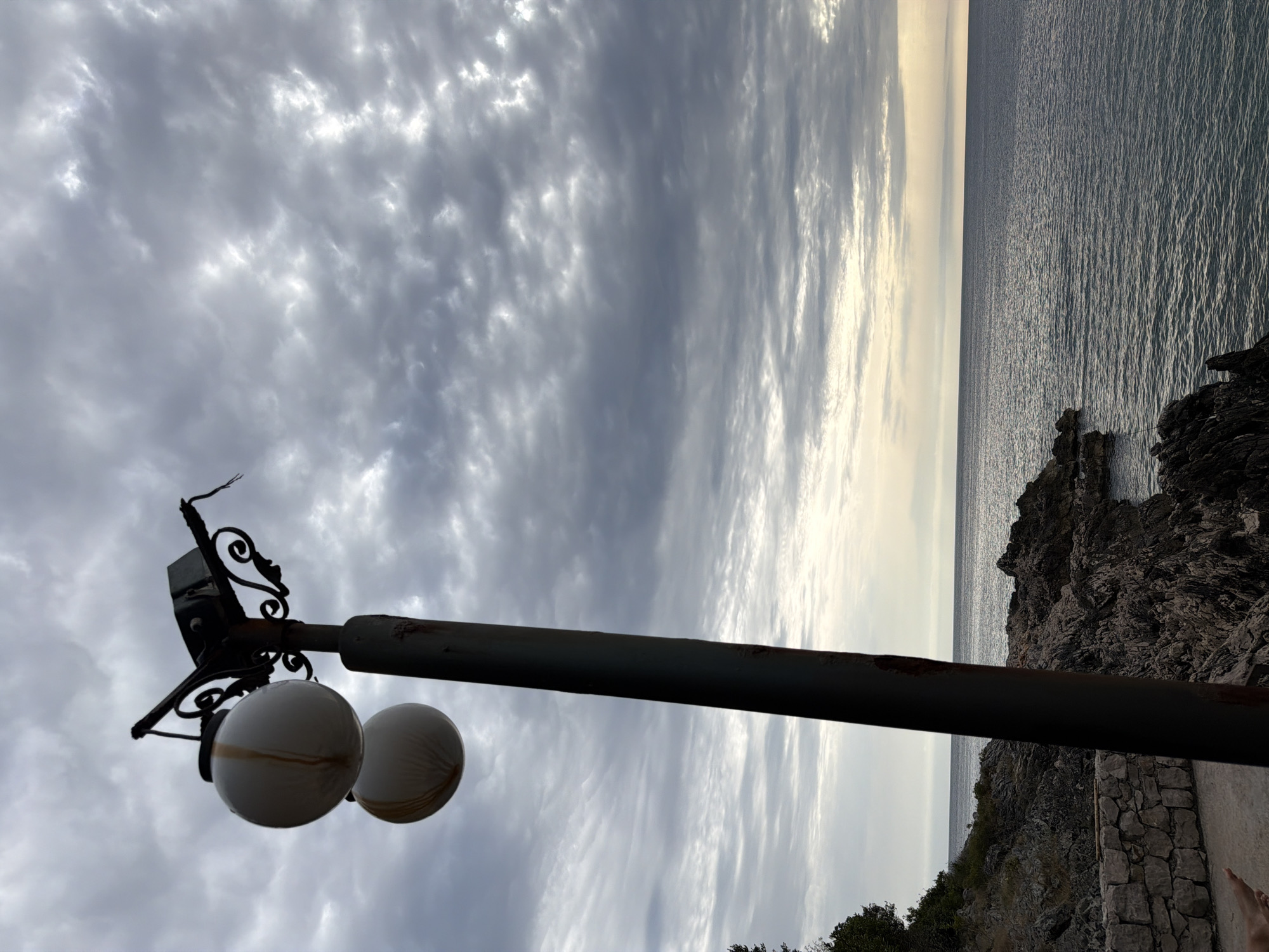 A tall, dark, weathered lamp post stands prominently in the foreground, its pole showing rust and a green patina. Two round, white globes hang from ornate black metalwork at the top, one slightly above the other. The upper globe has streaks of brown discoloration. The lamp post is positioned against a dramatic sky filled with heavy, gray clouds that are breaking in places to reveal lighter patches, suggesting a moody, overcast day.
In the background, a rocky coastline stretches out to the right, with dark, irregular rocks meeting the calm, dark blue sea. The water appears rippled, reflecting the overcast sky. To the left, the coastline is covered with dense green vegetation and a low stone wall borders a paved area in the immediate foreground. The overall scene evokes a sense of a coastal location, possibly a park or promenade, in a natural setting. The image appears to be taken from a slightly low angle, emphasizing the lamp post against the sky and sea. The location is specified as Kunje, Montenegro.