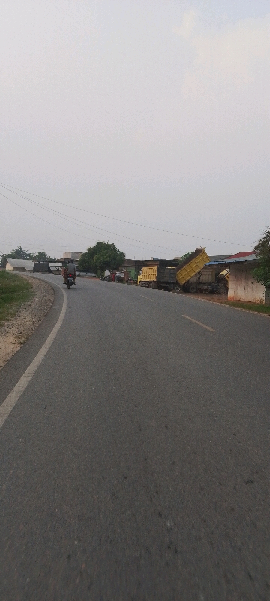 In Lenggadai Hilir, Indonesia, a dump truck is shown with its bed tilted upwards, indicating it is either being loaded or unloaded. The truck is parked on the side of a paved road, which curves to the left. Another dump truck is visible behind it. A motorcycle with two passengers is traveling away from the viewer on the road, heading towards the dump trucks. In the background, there are several low-rise buildings and trees. The sky is overcast, suggesting it is either early morning or late afternoon.